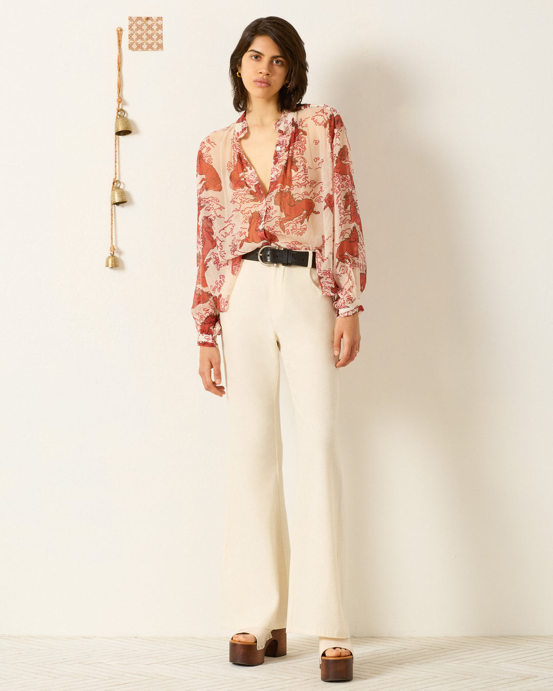 A woman stands against a white wall in the Poet Rouge Pegasus Blouse, paired with cream high-waisted pants, a black belt, and brown platform sandals. Small bells hang on the wall beside her.