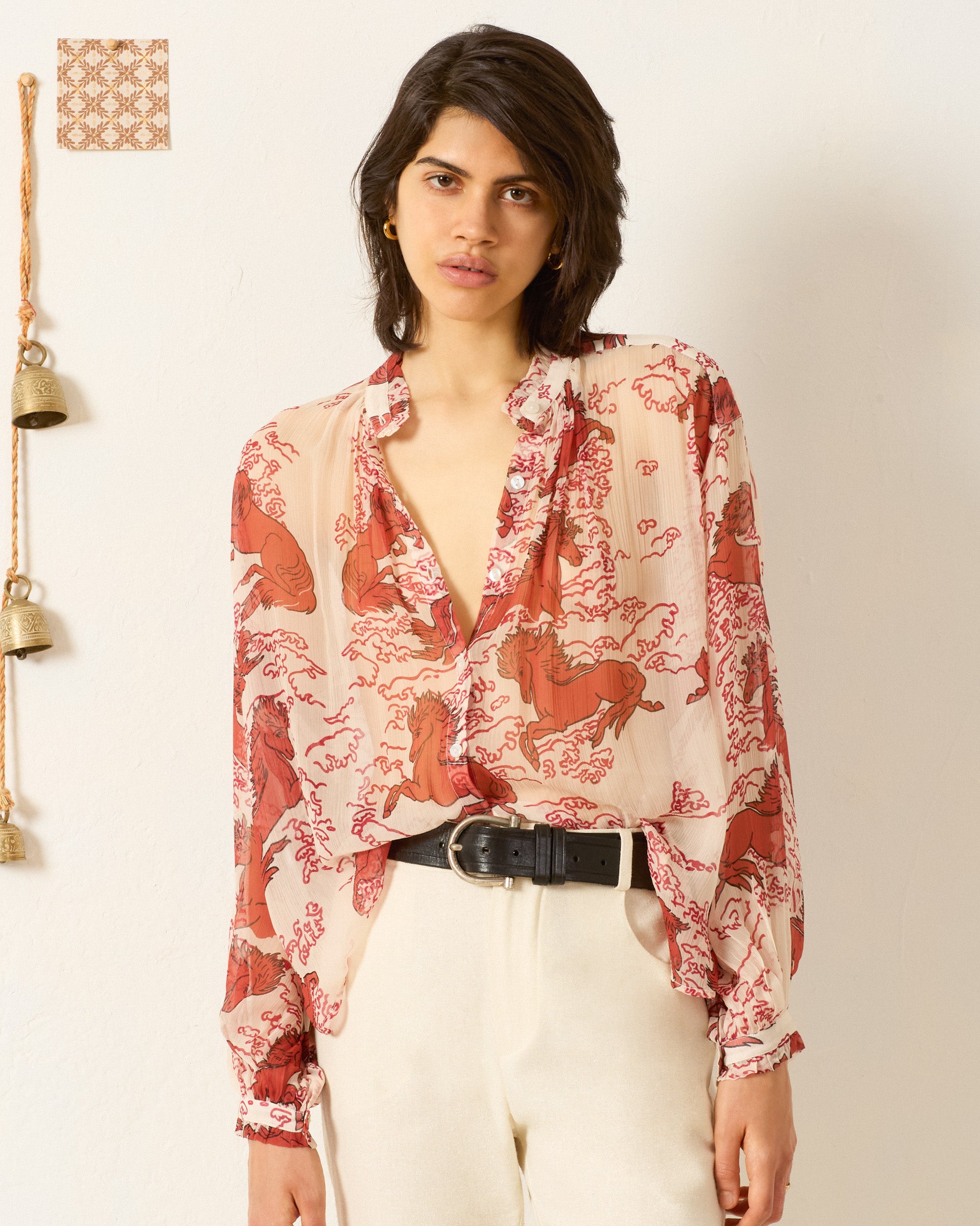 A woman stands indoors wearing the Poet Rouge Pegasus Blouse with a ruffled collar and red designs, paired with light high-waisted pants and a black belt; bells and a patterned square decorate the wall behind her.
