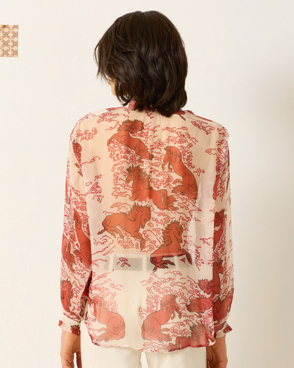 Person with short dark hair, seen from behind against a light background, wears the Poet Rouge Pegasus Blouse featuring a sheer cupro-chiffon fabric and a reddish mythical horse motif.