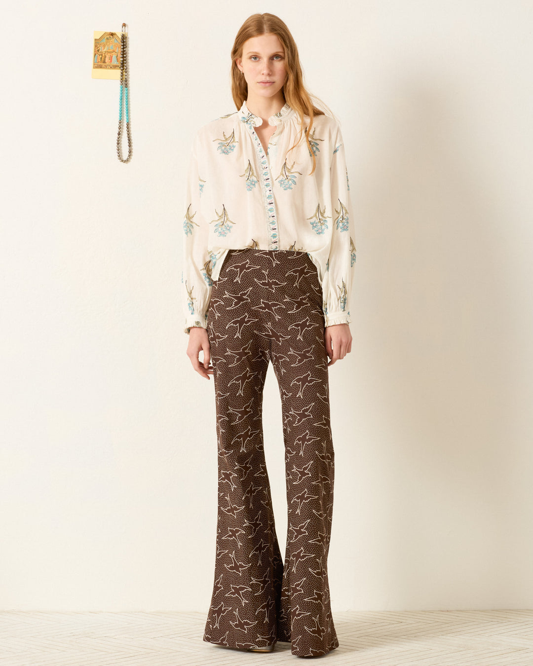 A woman stands against a plain white wall, wearing the Poet Sky Mist Flower Blouse and brown high-waisted wide-leg pants with a white bird pattern. A wall decoration hangs to her left.