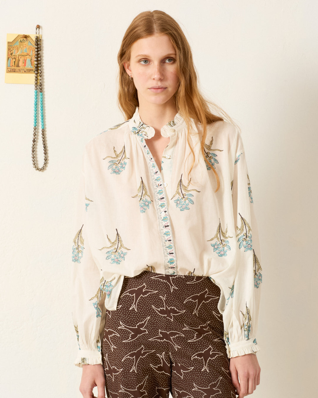 A woman stands against a white wall, wearing the Poet Sky Mist Flower Blouse with a ruffled collar and dark bird-print pants. A beaded necklace and a small card hang on the wall beside her.