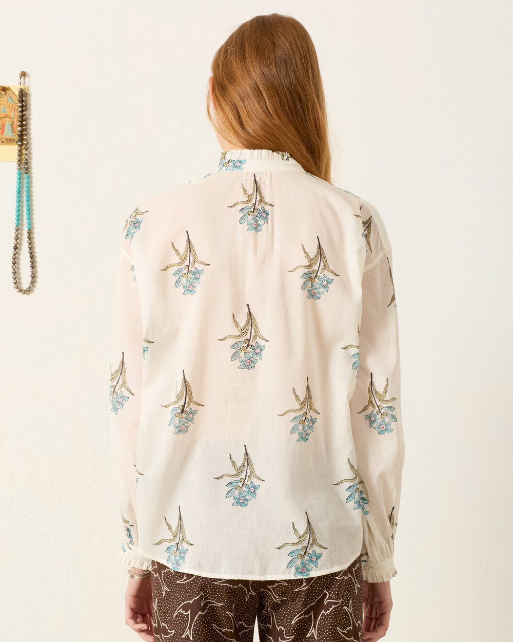 A person with long hair stands facing away, wearing the Poet Sky Mist Flower Blouse—a sheer, hand-block-printed cotton voile shirt with a floral pattern—and dark pants with a white abstract design.