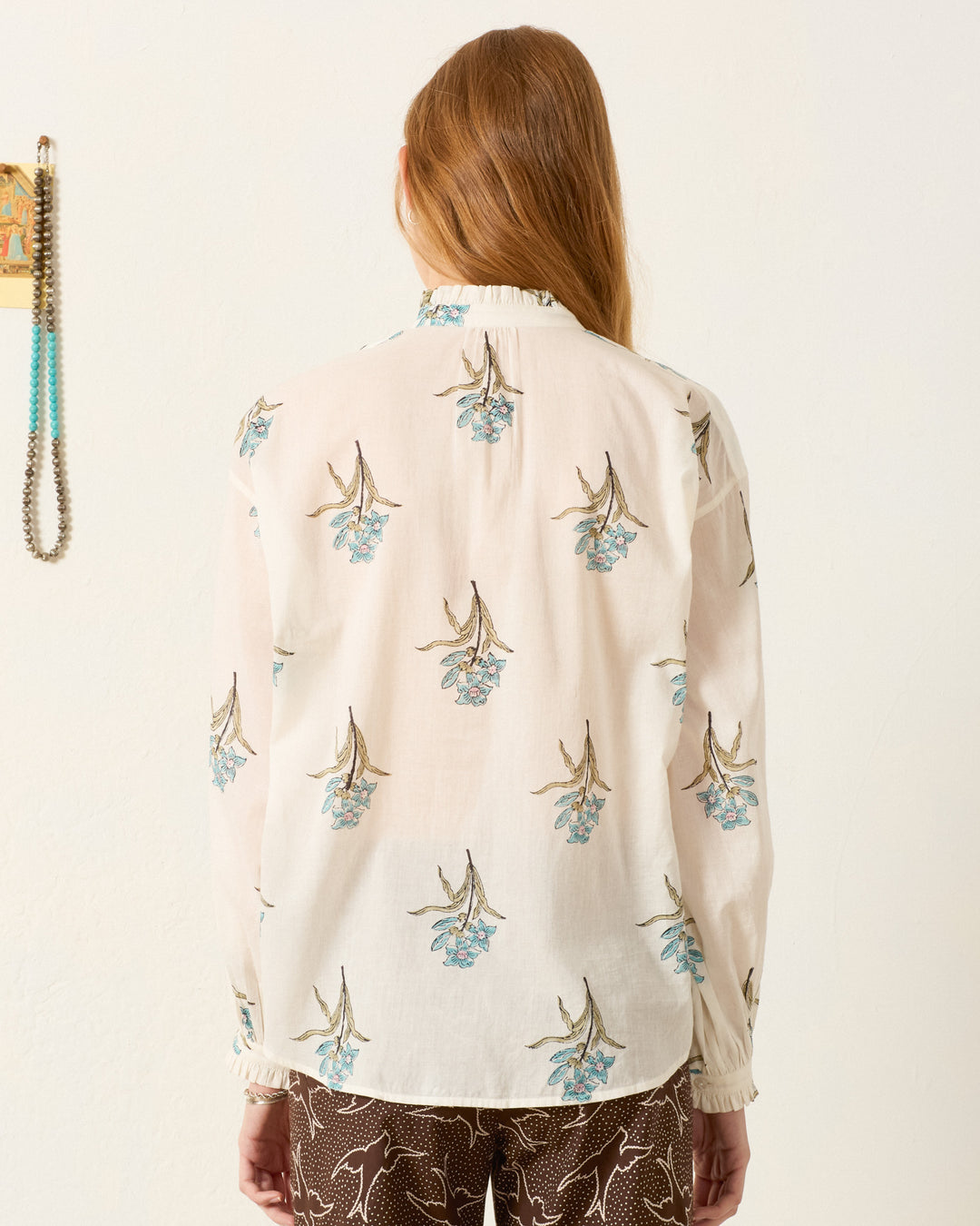 A person with long hair stands facing away, wearing the Poet Sky Mist Flower Blouse—a sheer, hand-block-printed cotton voile shirt with a floral pattern—and dark pants with a white abstract design.