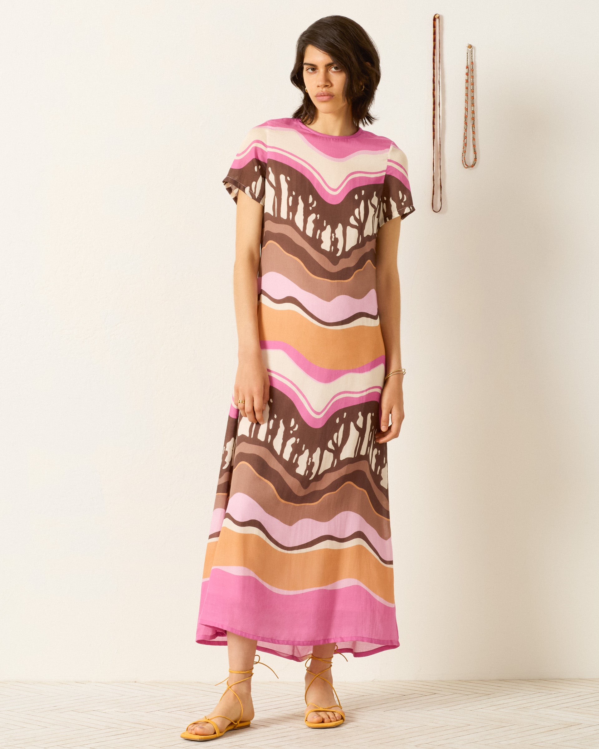 A person wears the Preeti Bloomshade Dress—a pink, brown, and tan abstract print A-line style—indoors against a plain wall, with sandals and two belts hanging nearby.