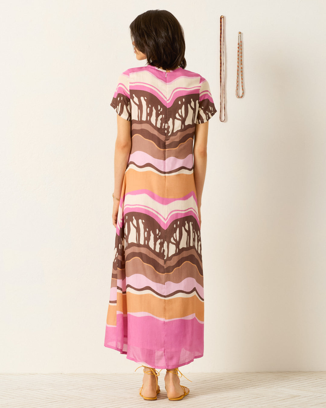 A person stands facing away, wearing the Preeti Bloomshade Dress—an ankle-length A-line dress with a wavy brown, pink, and tan abstract print—near a white wall with two necklaces hanging.