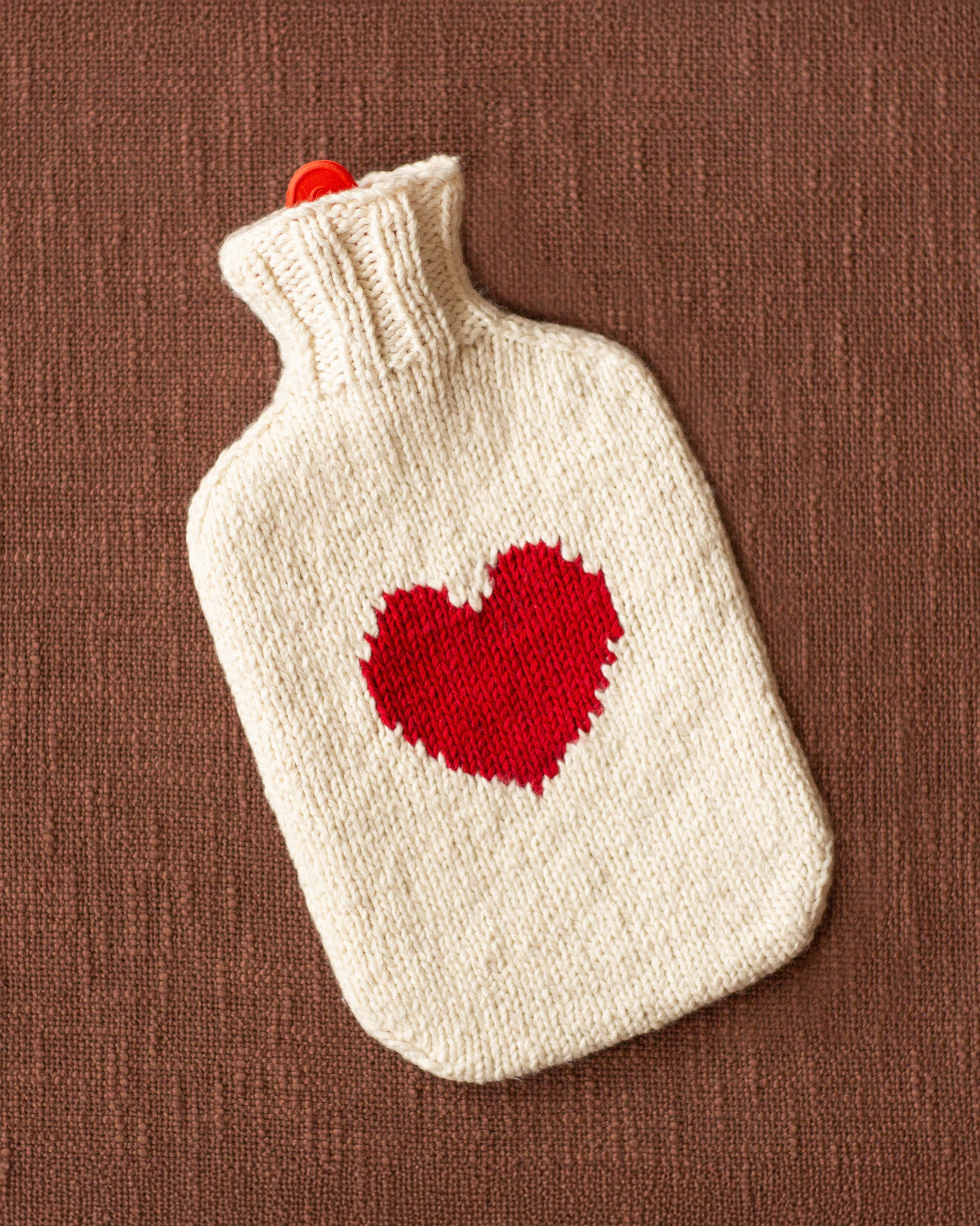 The Rio Heart Bottlecover is a cream-colored, hand-knitted hot water bottle cover with a red heart, shown on a brown textured surface—an ideal cozy holiday gift.