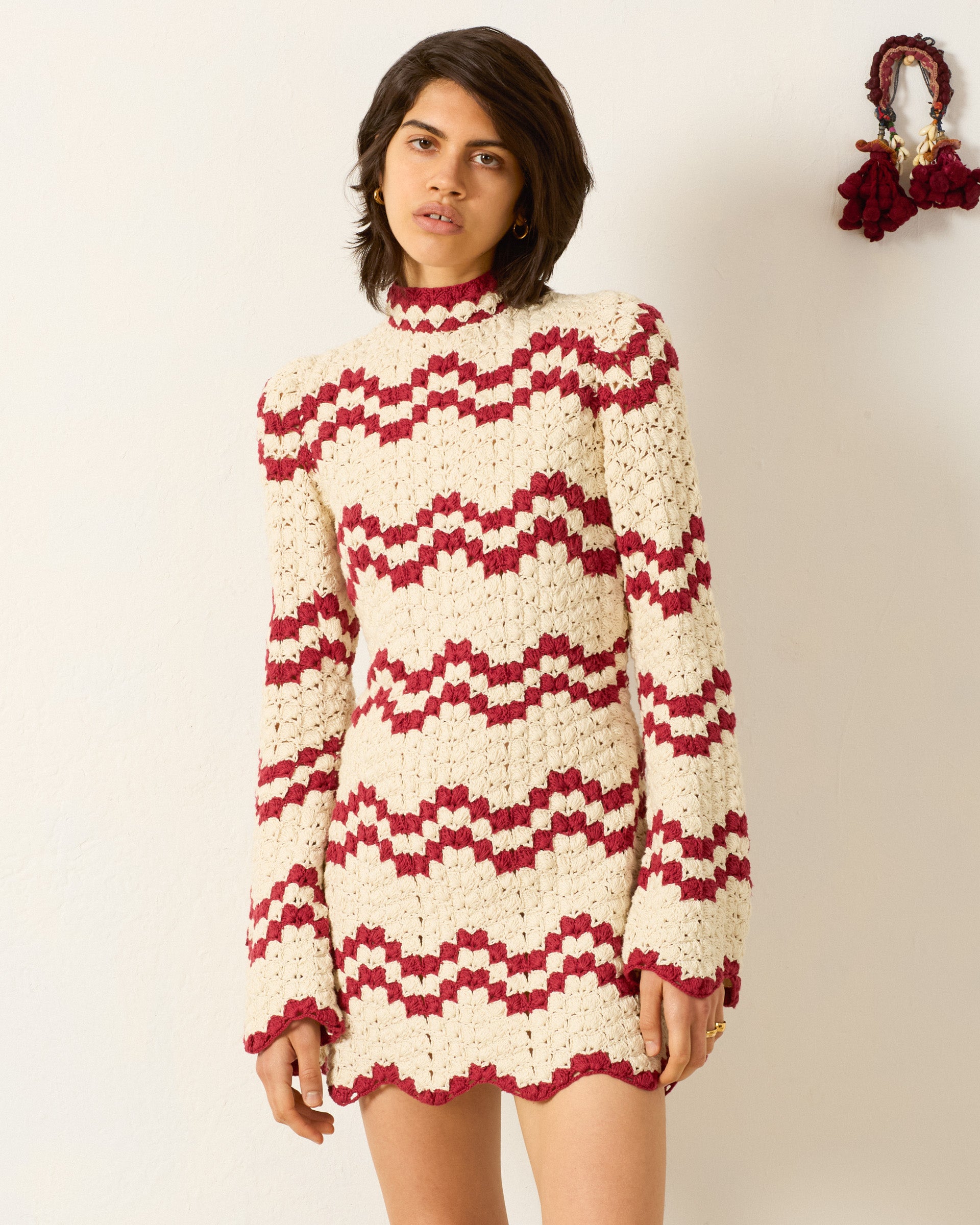 A woman stands against a plain wall wearing the Romy Ruby Stripe Dress, a long-sleeve, high-neck hand crocheted piece featuring cream and red zigzag stripes with a delicate scalloped hem.