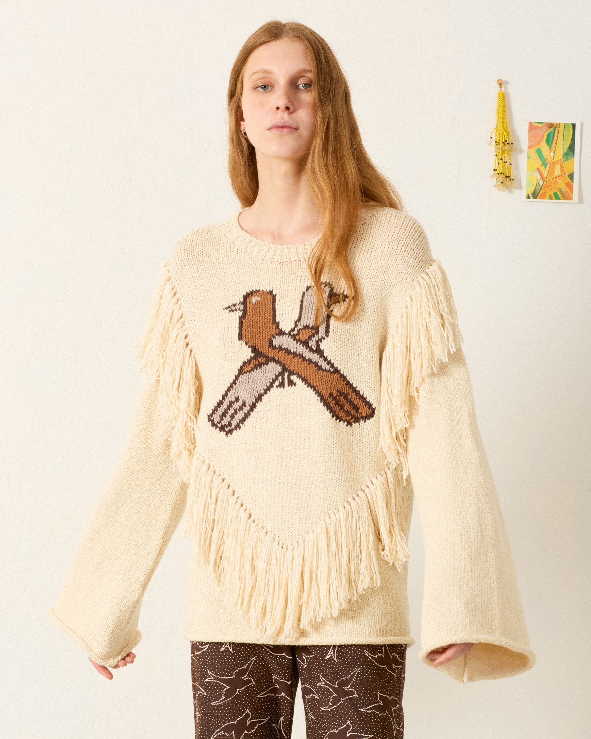 Woman stands by a white wall in the Rosa Lovebird Knit Sweater, featuring cozy baby alpaca, fringe, and a bird graphic, paired with dark patterned pants. Tassel and art hang behind her—ideal for winter layering.