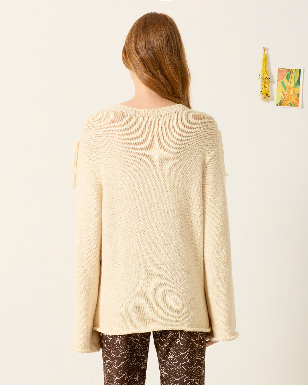 A person with long, light brown hair faces away from the camera, wearing the Rosa Lovebird Knit Sweater—a loose, cream-colored baby alpaca piece—paired with brown pants featuring a white bird pattern, against a plain wall.