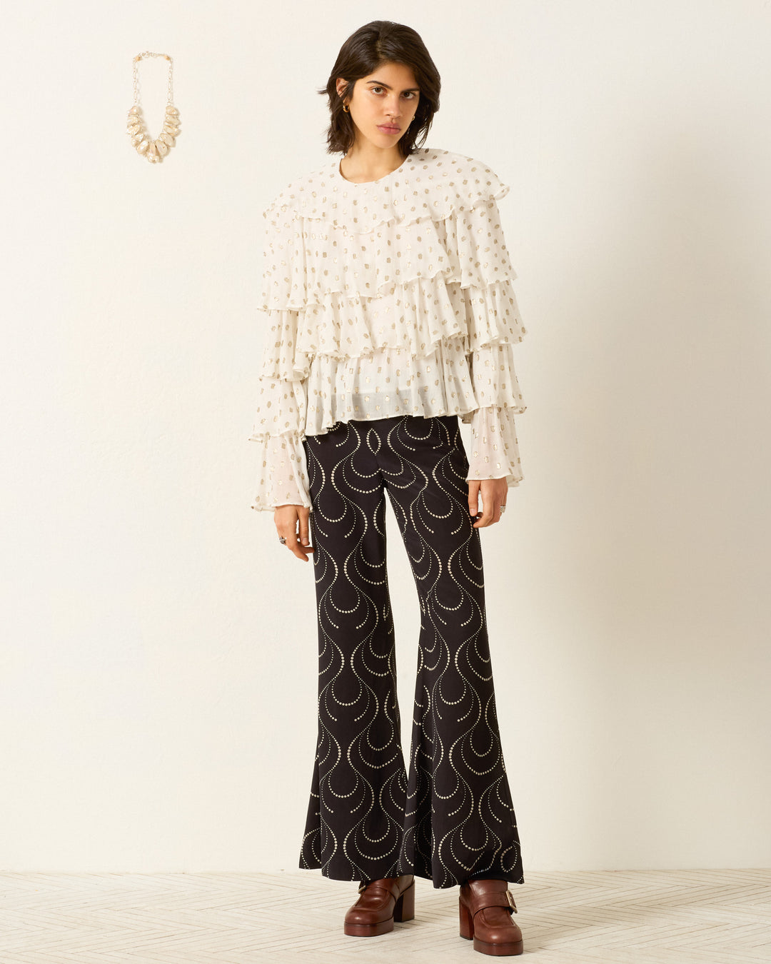 A person stands indoors wearing the Rufus Bronze Mist Top, black flared pants with a wavy pattern, and brown platform shoes; a beaded necklace hangs on the wall.