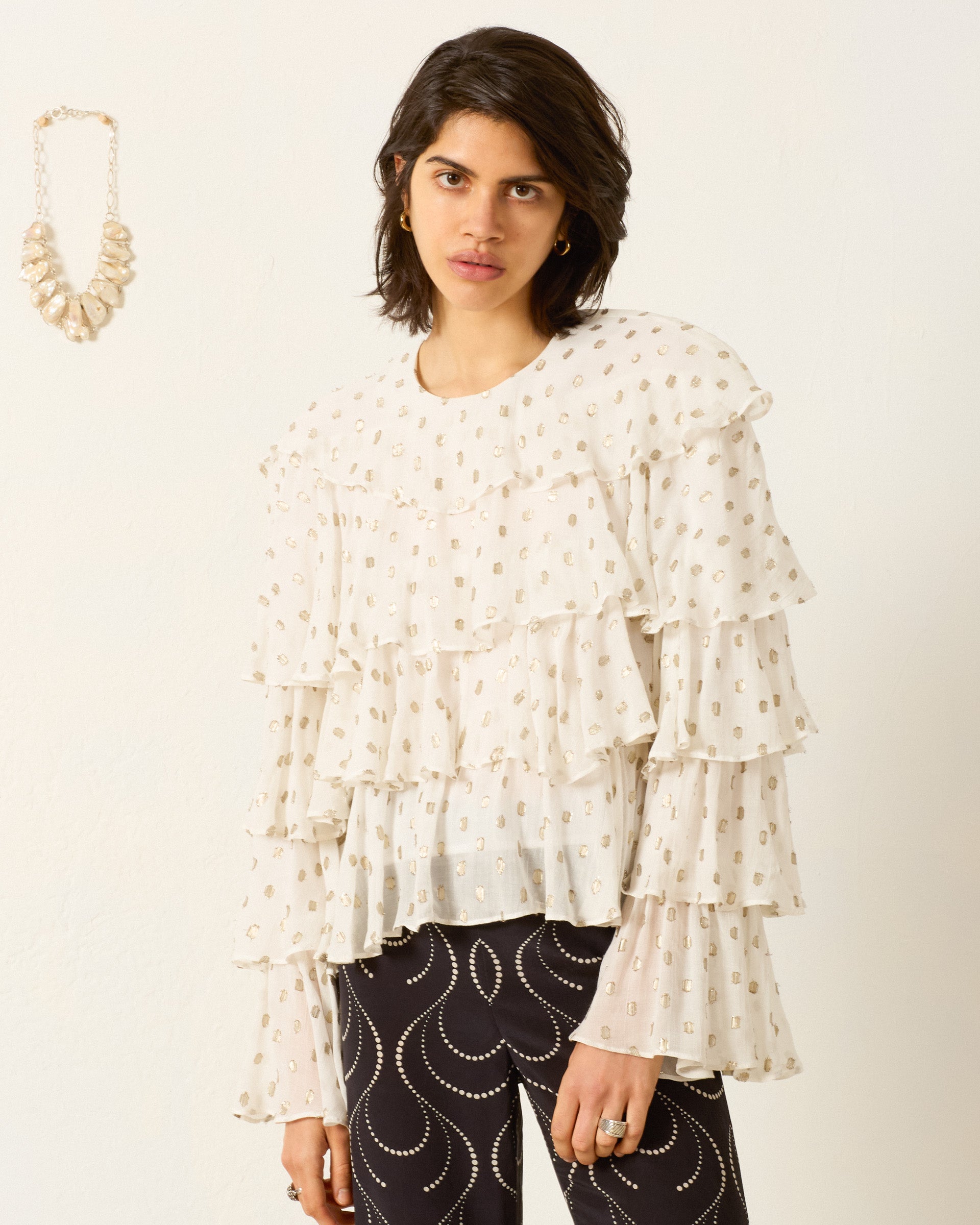 Indoors, a woman wears the Rufus Bronze Mist Top—a white ruffle blouse with gold patterns—paired with black pants featuring white curved lines. A beaded necklace is displayed on the wall beside her.