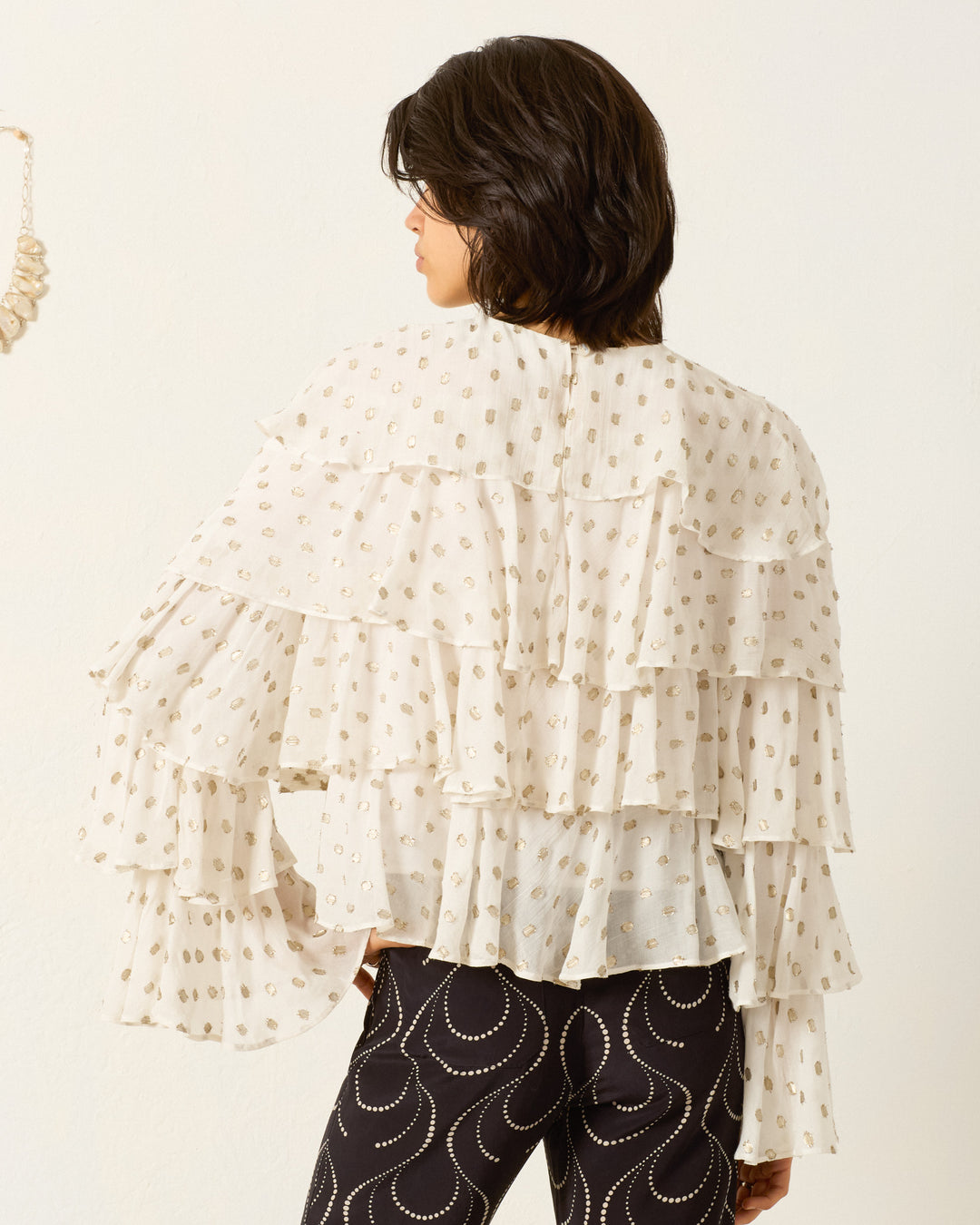 Person standing with back to camera, wearing the Rufus Bronze Mist Top— a white ruffle blouse with polka dots— paired with black pants featuring bold white swirl patterns.