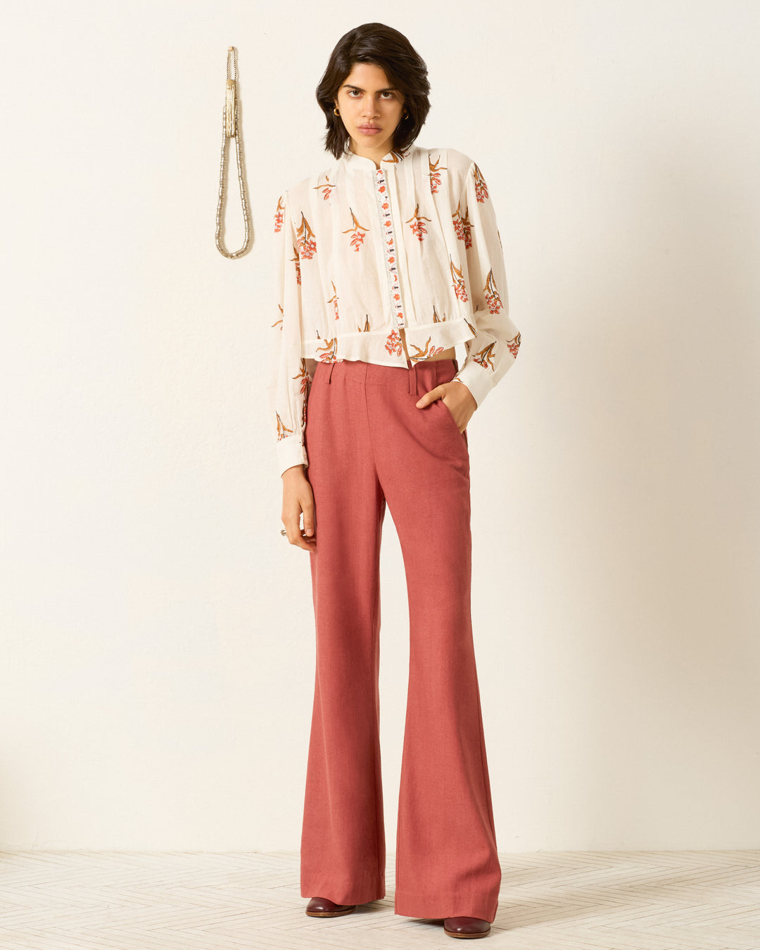 A person stands indoors wearing the Ryland Persimmon Flower Shirt, a hand-block-printed cotton voile blouse with a red floral pattern, paired with high-waisted, wide-leg red trousers. One hand is in their pocket.