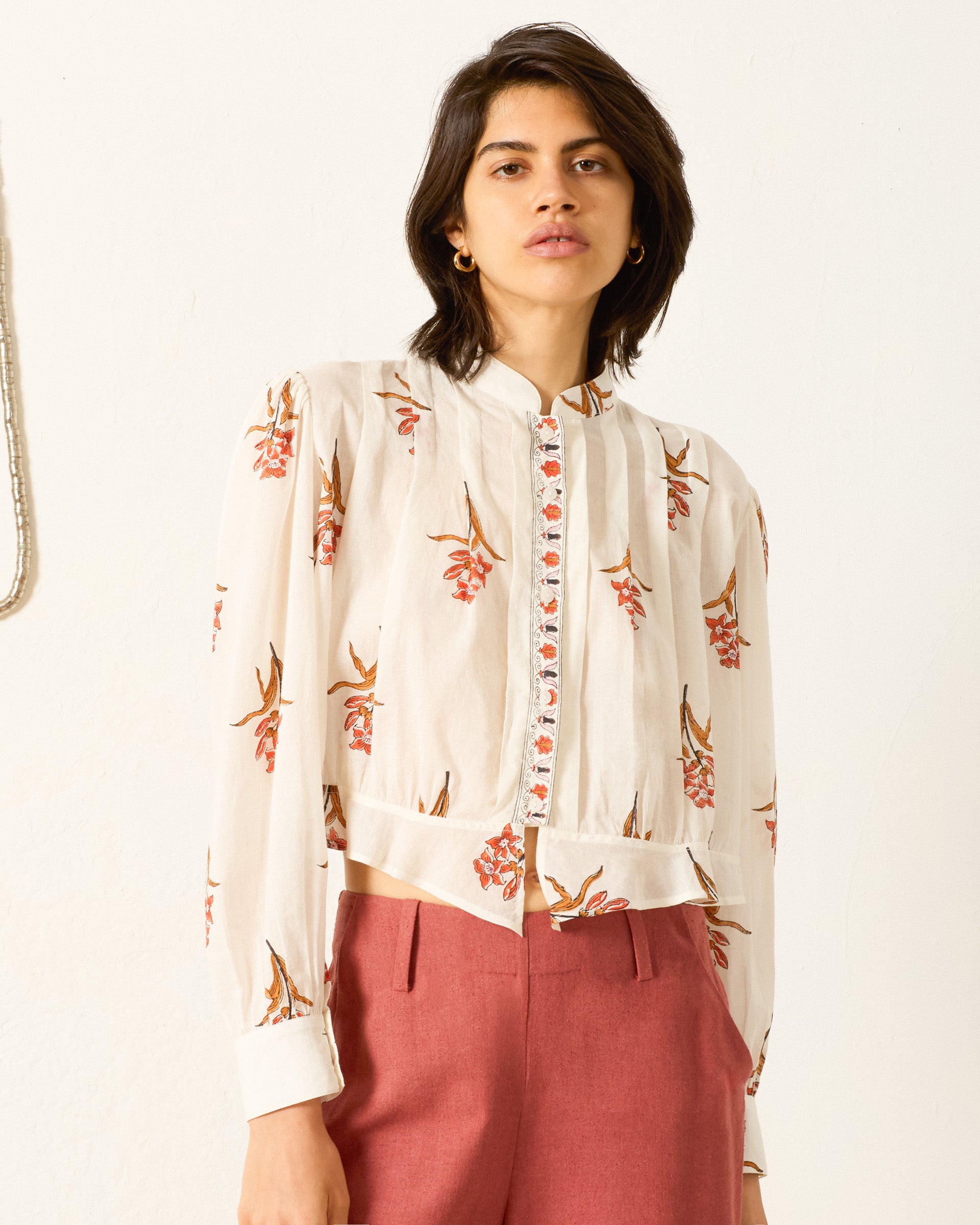 A woman stands against a plain background wearing the Ryland Persimmon Flower Shirt, a white, hand-block-printed cotton voile blouse with a floral print, paired with high-waisted pink trousers.