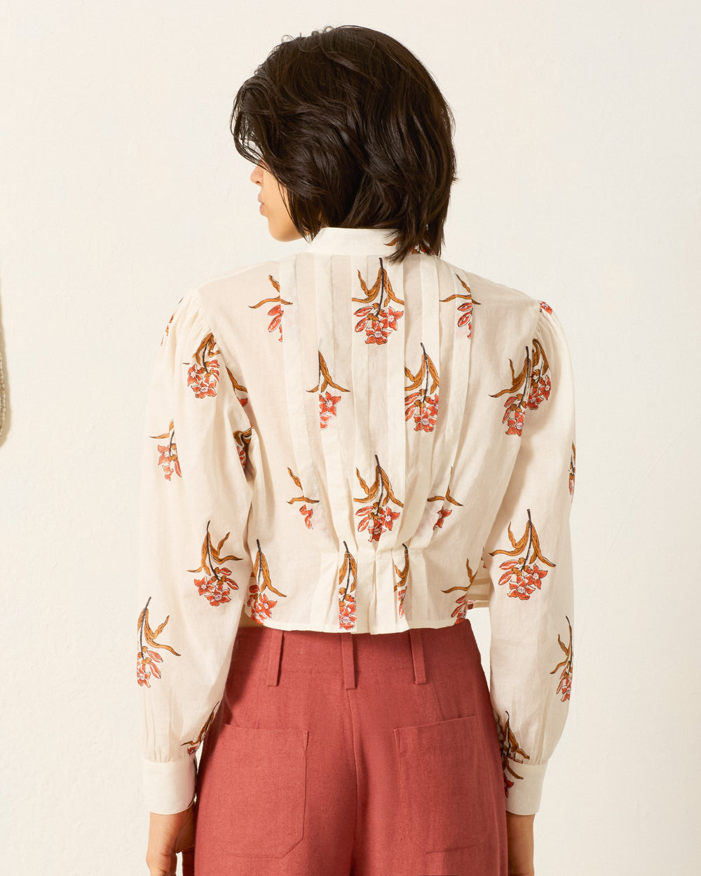 A person with short dark hair is shown from the back, wearing the Ryland Persimmon Flower Shirt and high-waisted rust-colored pants.