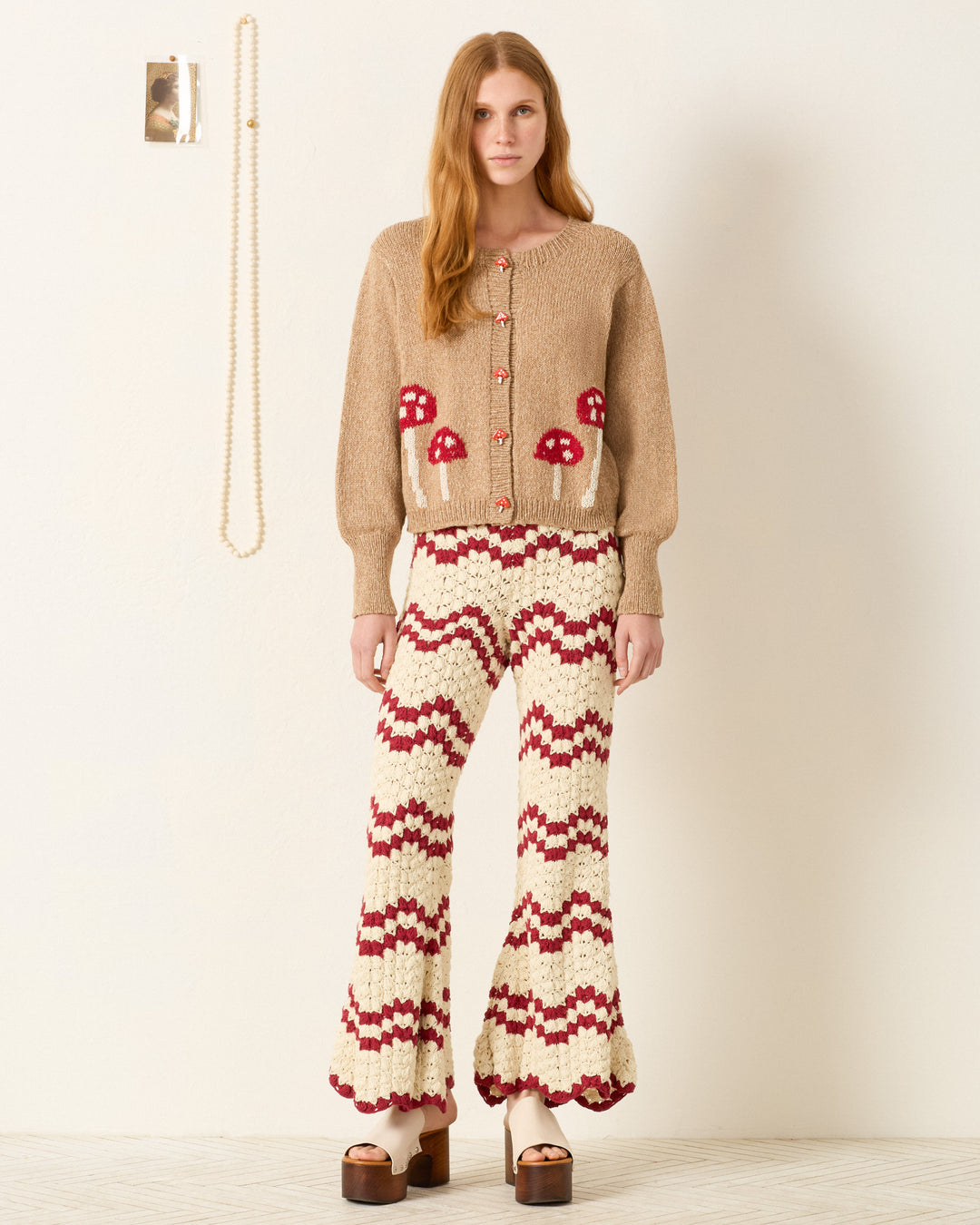 A woman stands against a plain wall, wearing the Sammi Mushroom Knit Cardigan and red-and-white zigzag crochet pants, paired with platform sandals.