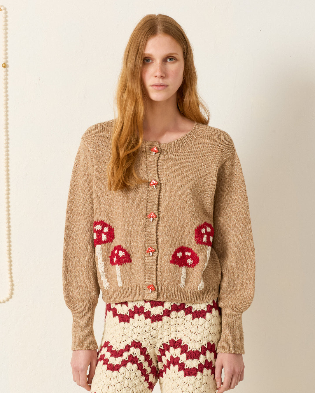 A woman with long red hair wears the Sammi Mushroom Knit Cardigan, featuring red and white toadstool motifs and handmade buttons, paired with patterned crochet pants, standing against a plain light-colored wall.