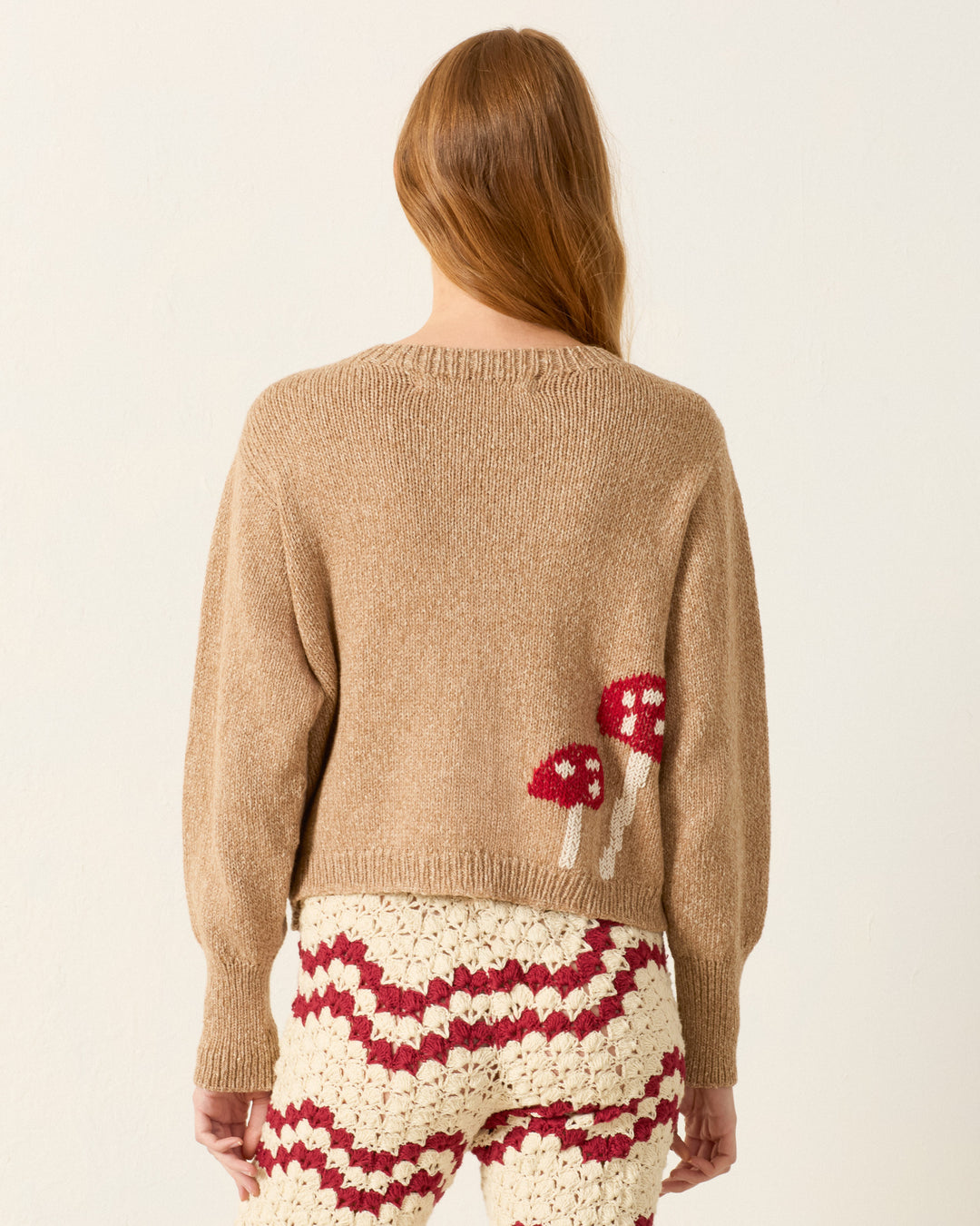 A person with long hair wears the Sammi Mushroom Knit Cardigan, a beige sweater with a toadstool motif on the back, paired with cream crochet pants featuring a red wavy pattern.