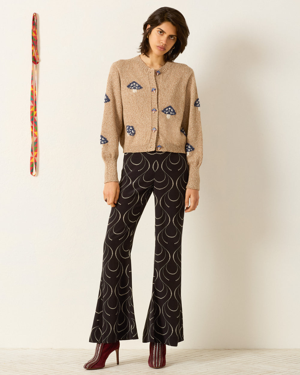 A woman stands indoors wearing the Sammi Champignon Knit Cardigan with blue patterns, black flared pants with white curved designs, and burgundy heeled boots. A colorful belt hangs on the wall beside her.