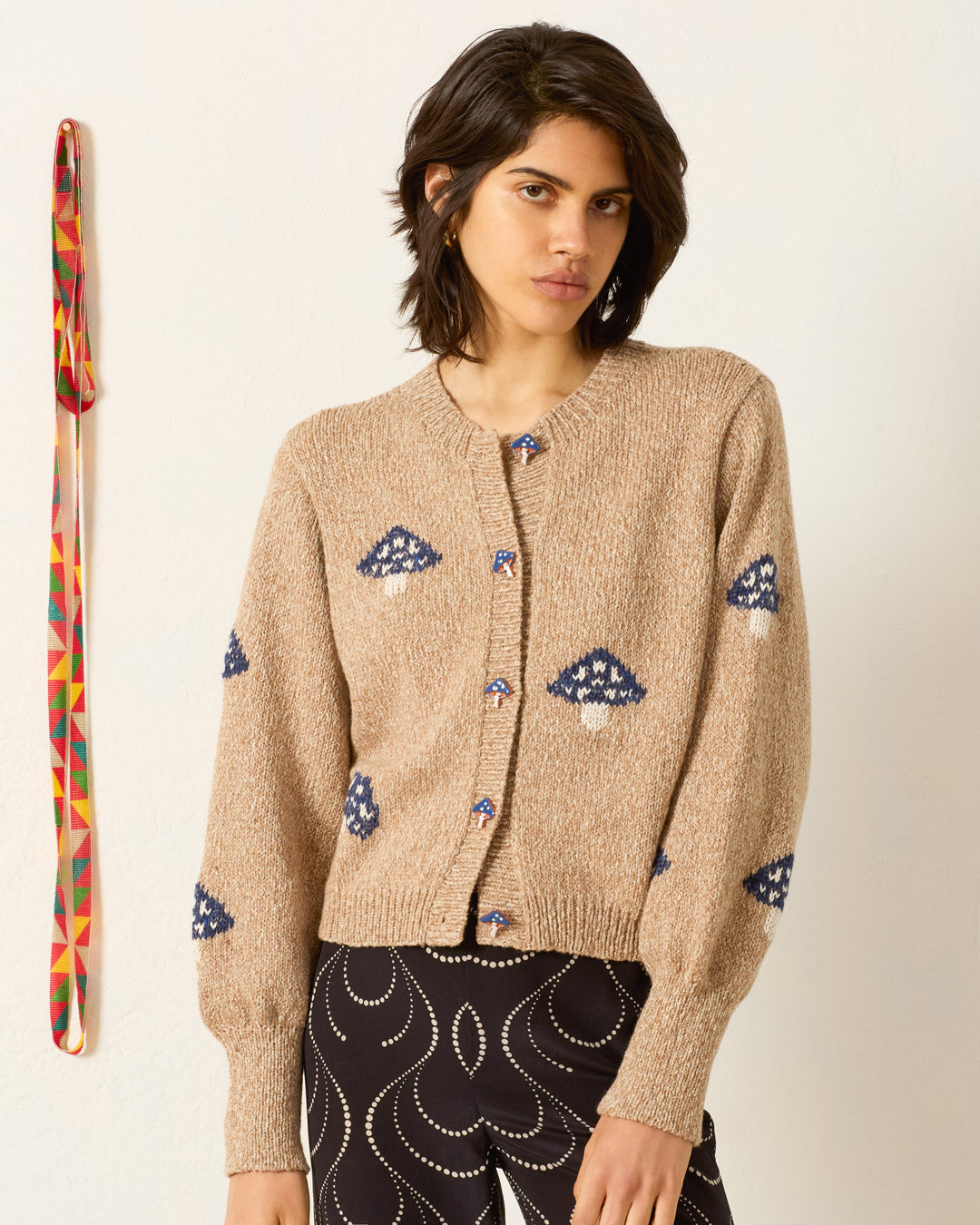 A woman wears the Sammi Champignon Knit Cardigan—a beige baby alpaca piece with blue and white mushroom motifs and handmade buttons—paired with black pants featuring a white swirl, standing before a plain wall with a colorful woven strap on the left.