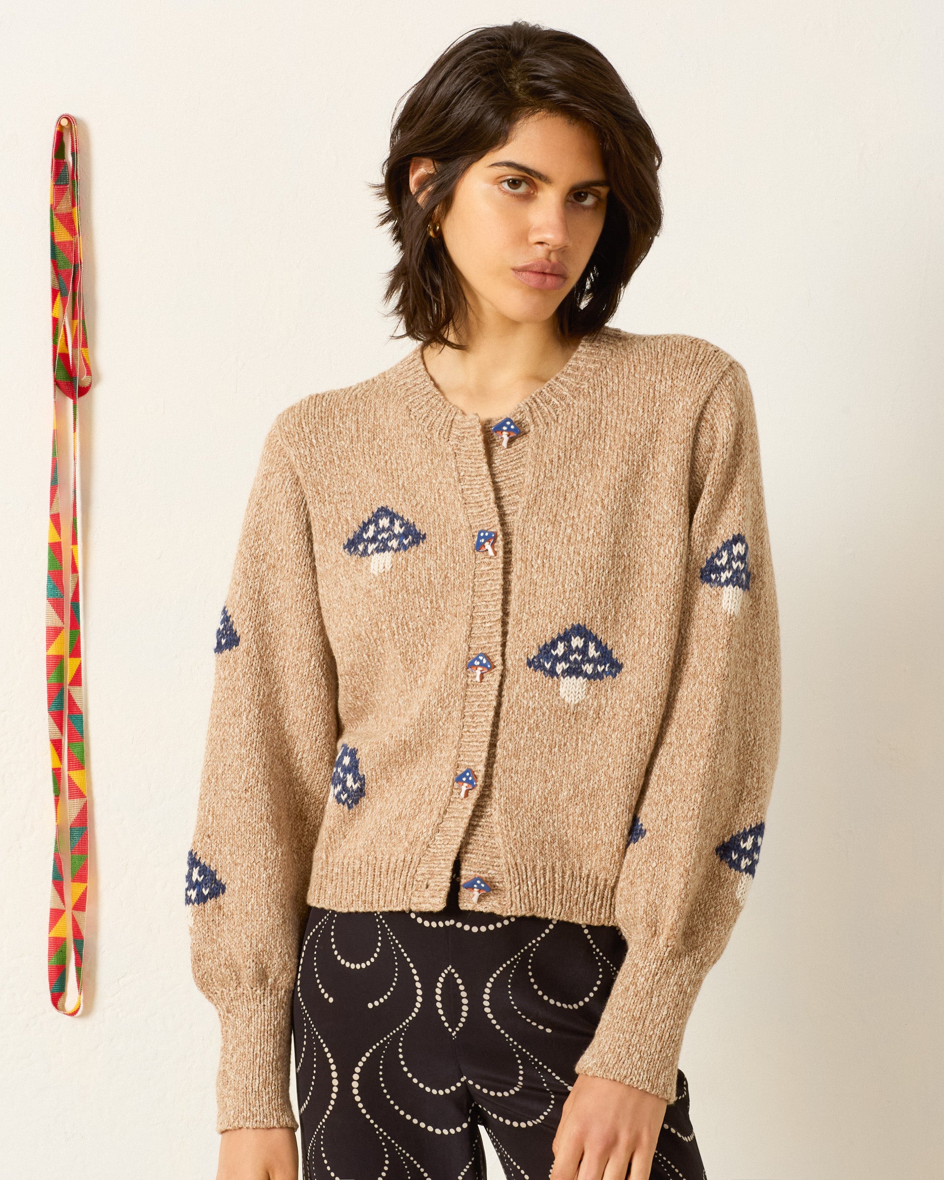 A woman wears the Sammi Champignon Knit Cardigan—a beige baby alpaca piece with blue and white mushroom motifs and handmade buttons—paired with black pants featuring a white swirl, standing before a plain wall with a colorful woven strap on the left.