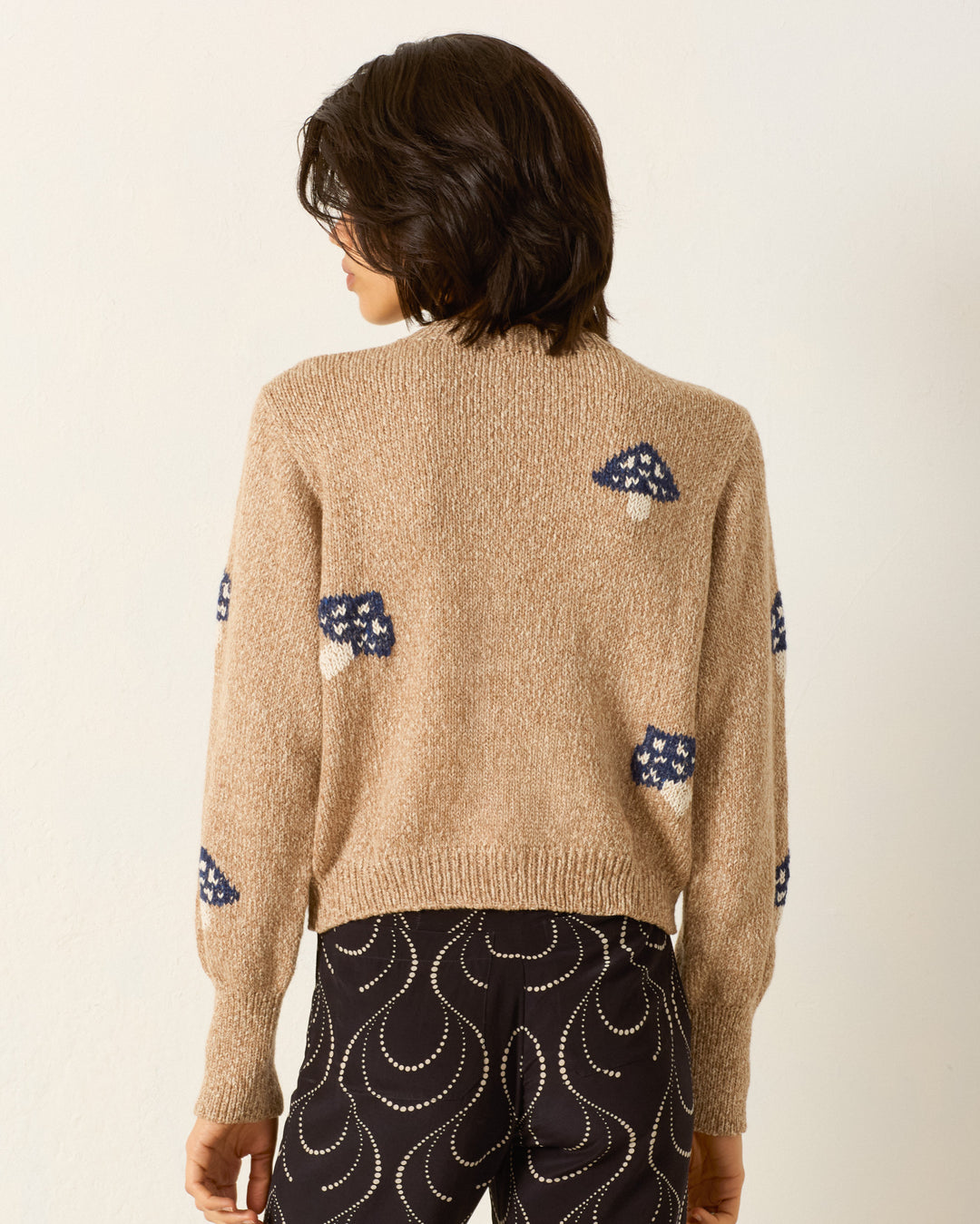 Person stands with their back to the camera, wearing the Sammi Champignon Knit Cardigan in beige with blue and white mushroom motifs, paired with dark pants decorated with white swirl designs.