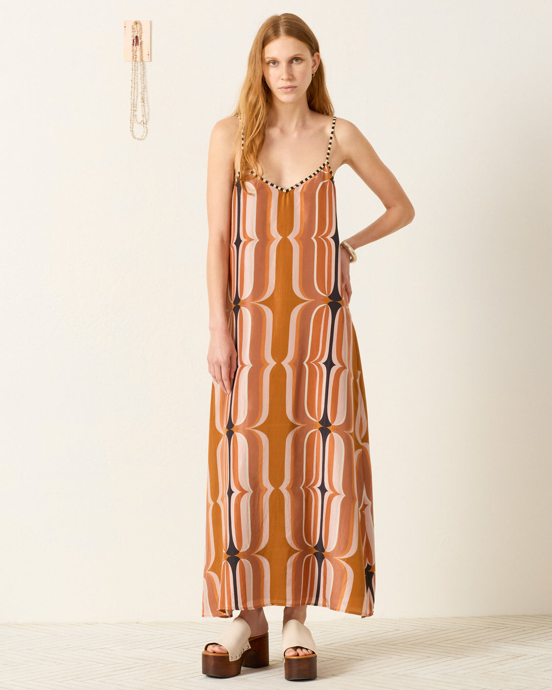A woman stands indoors wearing the Spice Market Mesa Verde Dress, a retro-inspired slip dress with a geometric brown and pink pattern, paired with platform sandals and minimal jewelry.