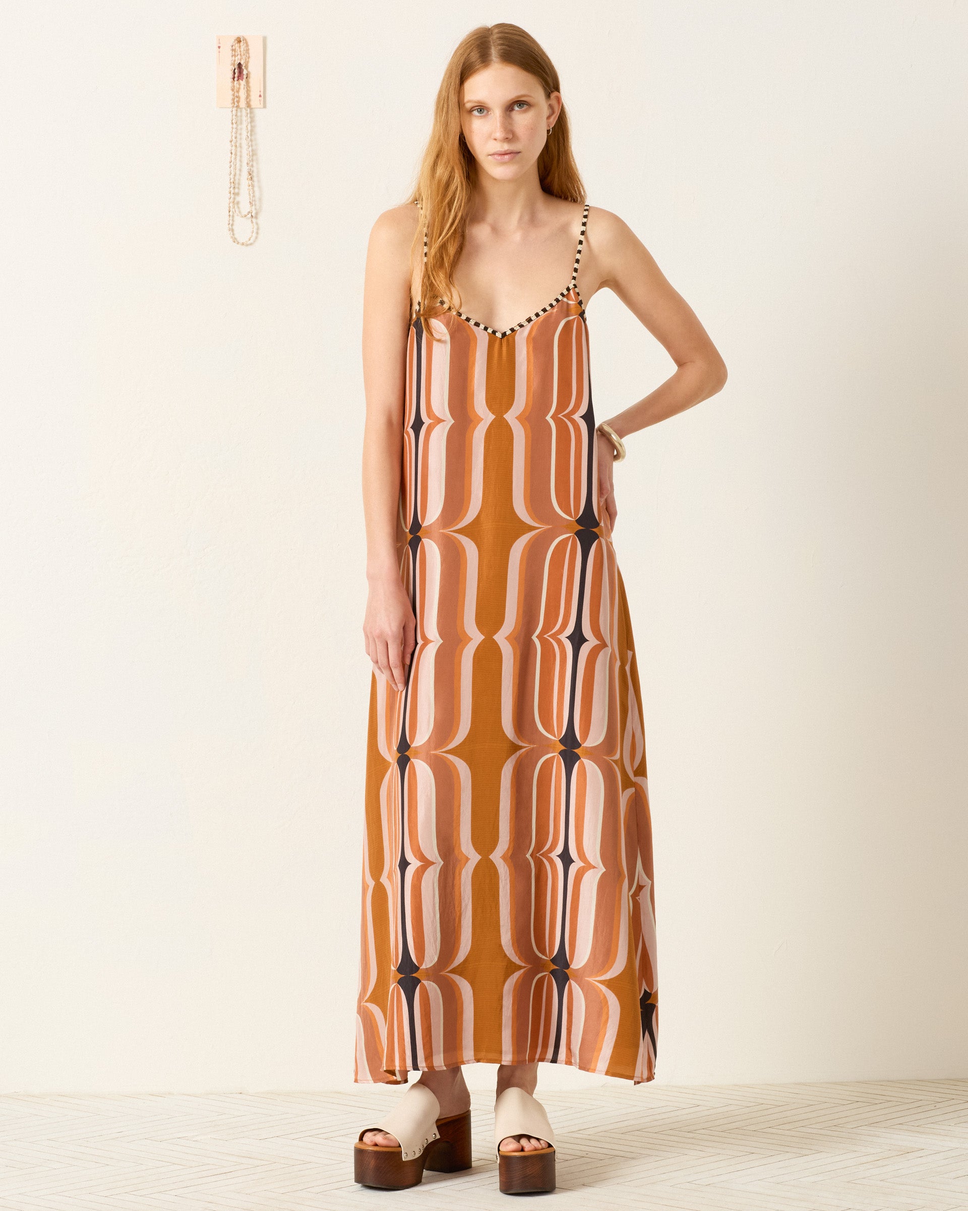 A woman stands indoors wearing the Spice Market Mesa Verde Dress, a retro-inspired slip dress with a geometric brown and pink pattern, paired with platform sandals and minimal jewelry.