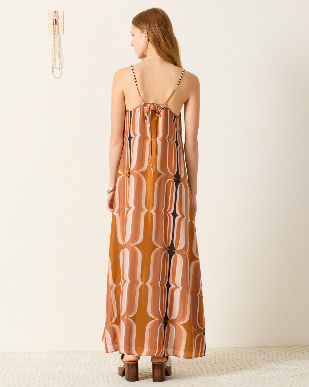 A woman faces away, wearing the Spice Market Mesa Verde Dress—a long, retro slip dress with an orange and white geometric pattern—and black platform sandals in a minimal, light-colored room.