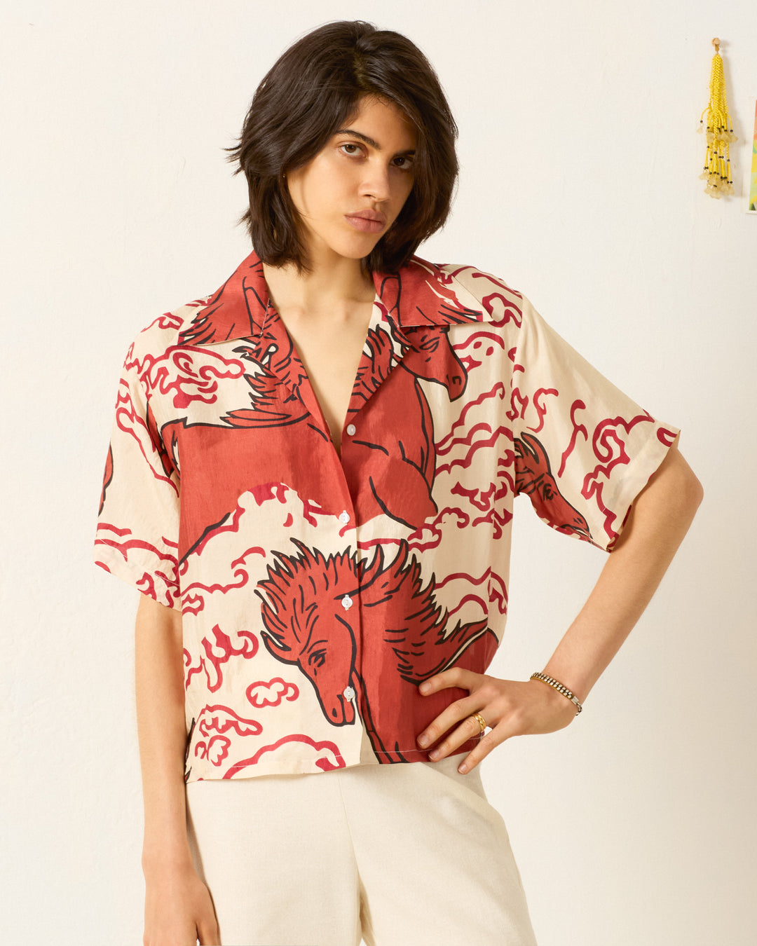 A person with shoulder-length hair wears the Stevie Rouge Pegasus Shirt—a cream-colored, short-sleeve, hand-loomed silk shirt with a red mythical horse motif—and light pants against a plain wall.