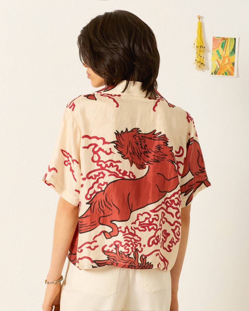 Person standing with their back to the camera, wearing the Stevie Rouge Pegasus Shirt—a beige hand-loomed silk shirt with a large red graphic. Small wall decorations can be seen in the background.