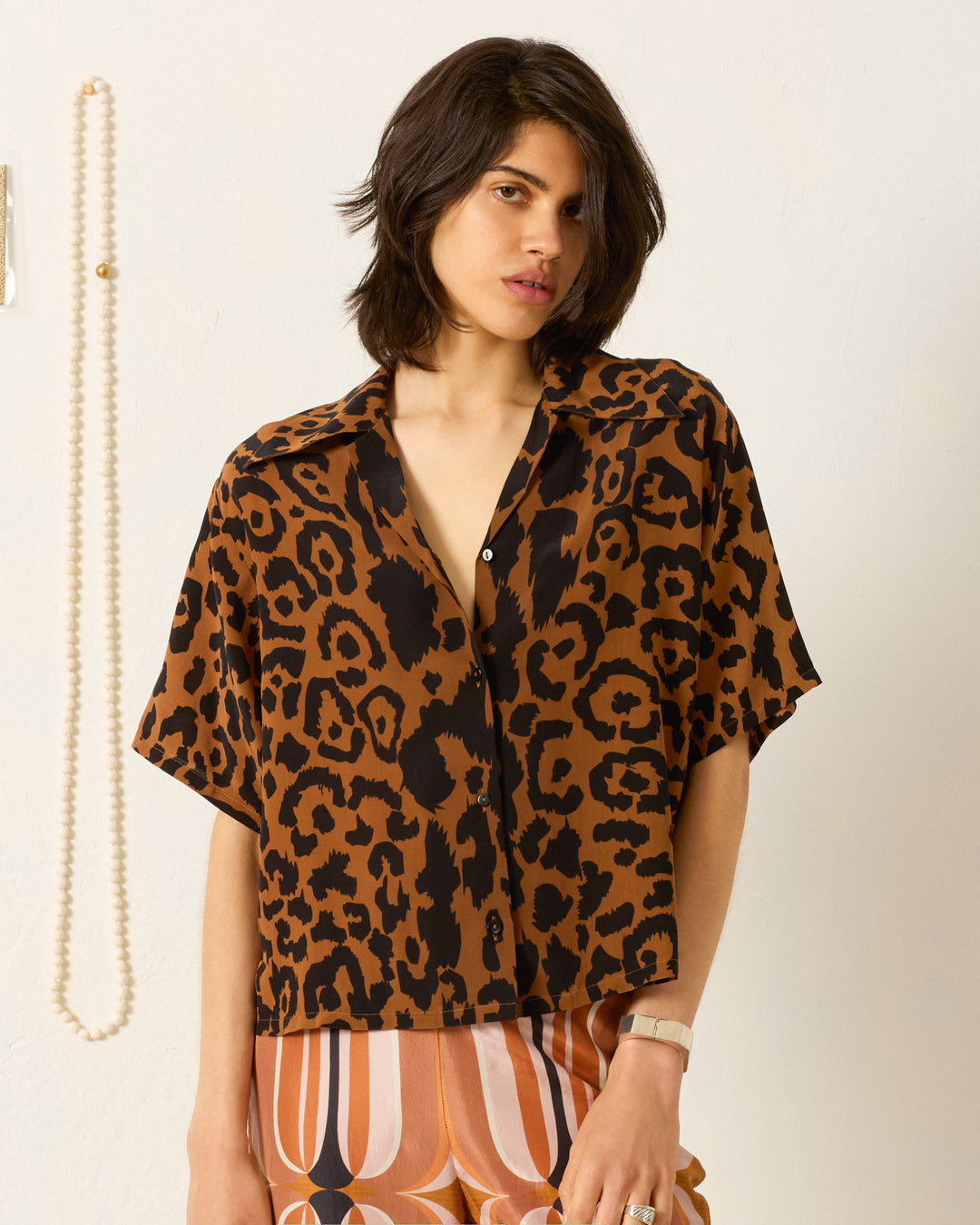 A person wears the Stevie Leopard Silk Shirt—loose, short-sleeved, and vintage-inspired in brown and black leopard print—paired with patterned pants, standing before a white wall with a hanging pearl necklace.