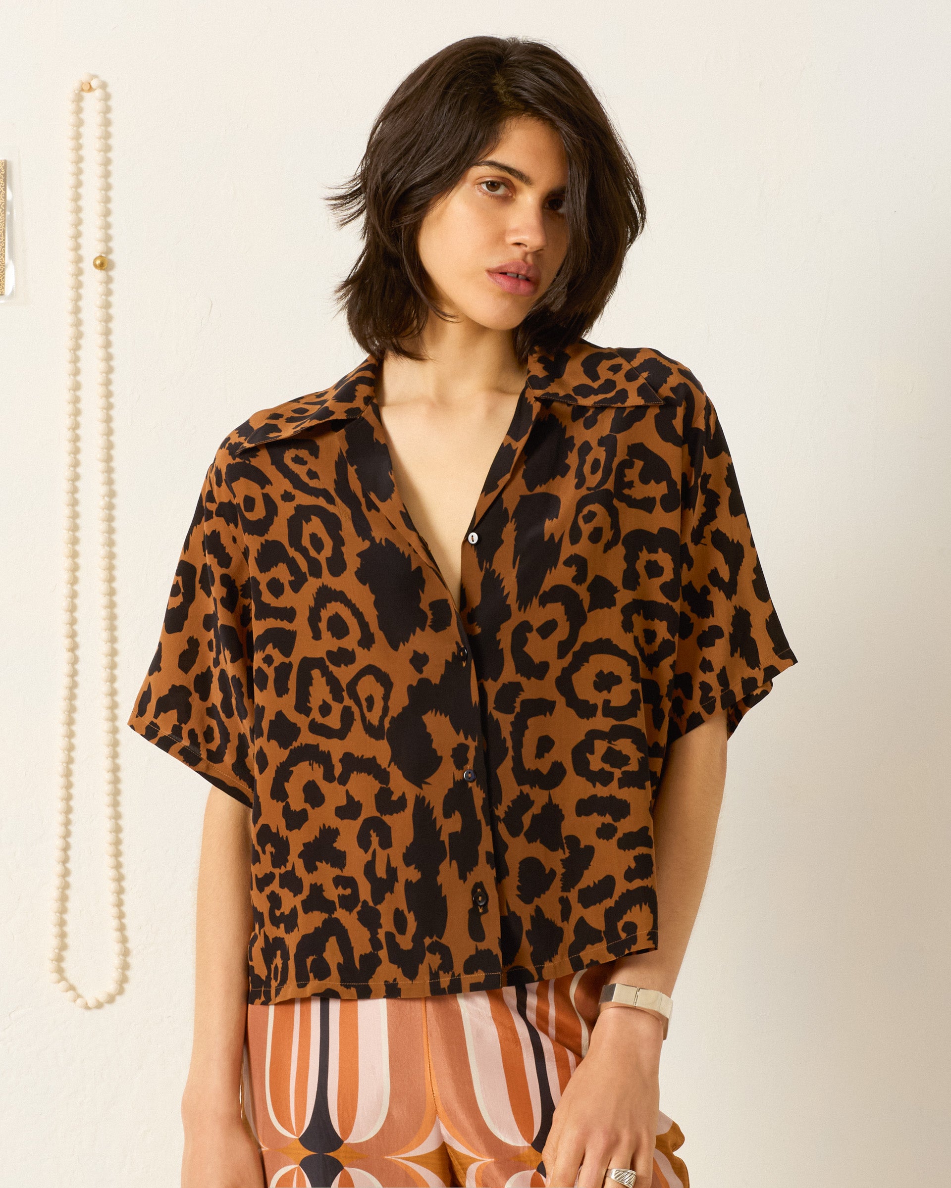 A person wears the Stevie Leopard Silk Shirt—loose, short-sleeved, and vintage-inspired in brown and black leopard print—paired with patterned pants, standing before a white wall with a hanging pearl necklace.