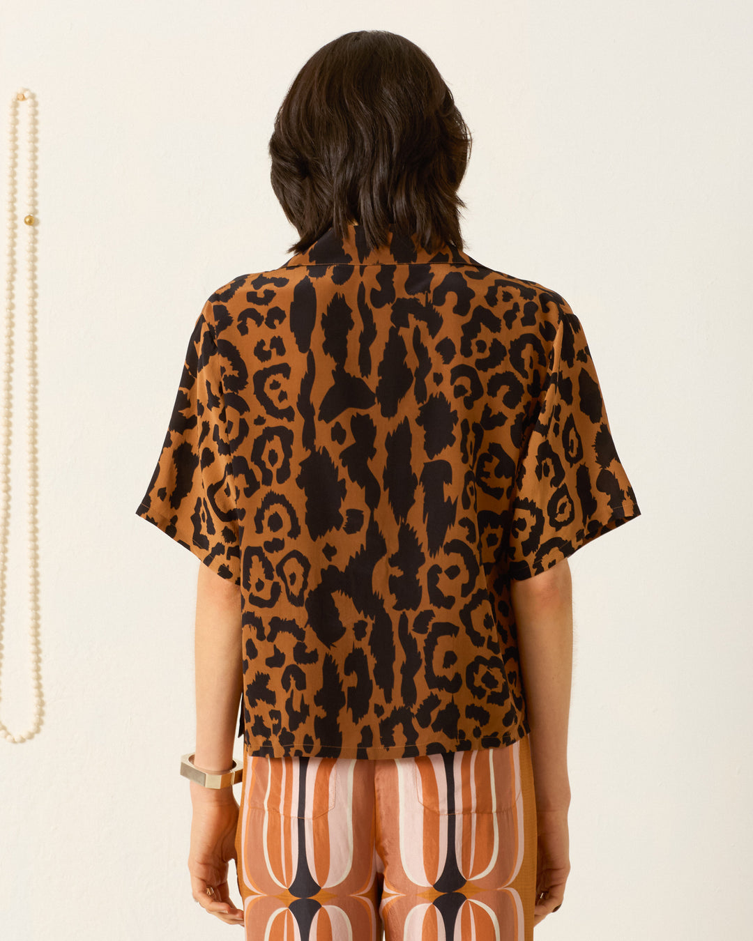 A person stands facing away, wearing the Stevie Leopard Silk Shirt—short-sleeved and vintage with a brown leopard print—paired with patterned pants featuring brown, orange, and white vertical designs.