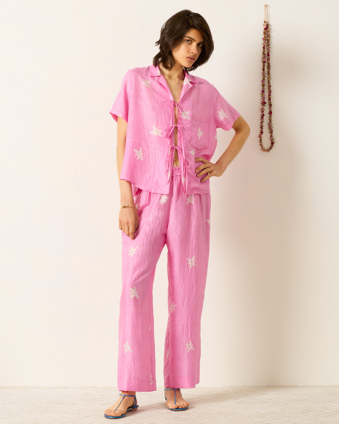 A person stands indoors near a garland-wrapped wall, wearing the Styla Pink Jasmine Shirt—a loose-fitting, hand-block-printed pink two-piece with white floral patterns.