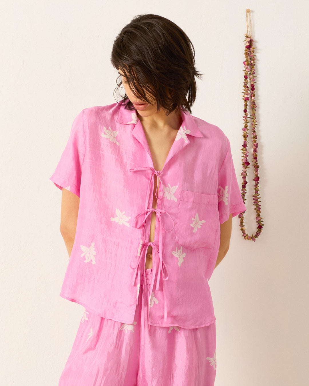 A person wearing the Styla Pink Jasmine Shirt, a loose hand-block-printed floral top with front tie closures, paired with matching pink shorts, stands against a plain wall adorned with a floral garland.