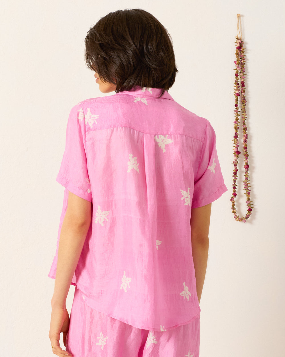 A person with short dark hair, seen from behind, wears the Styla Pink Jasmine Shirt— a loose, short-sleeved top featuring pink and white floral patterns. A string of dried flowers is displayed on the wall.