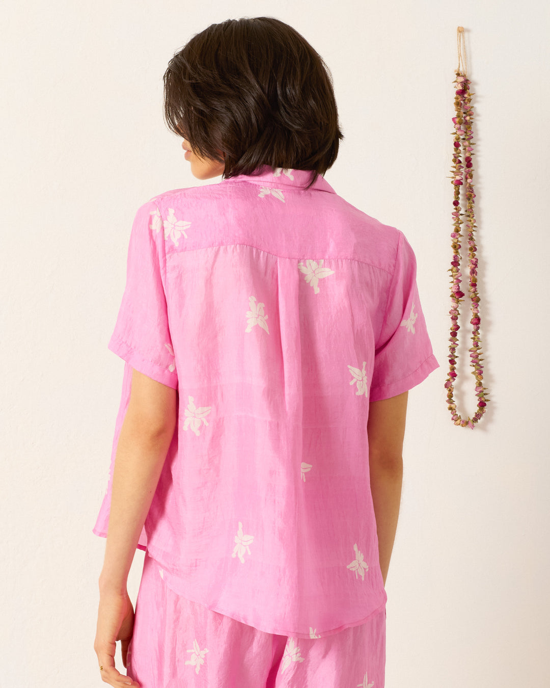 A person with short dark hair, seen from behind, wears the Styla Pink Jasmine Shirt— a loose, short-sleeved top featuring pink and white floral patterns. A string of dried flowers is displayed on the wall.