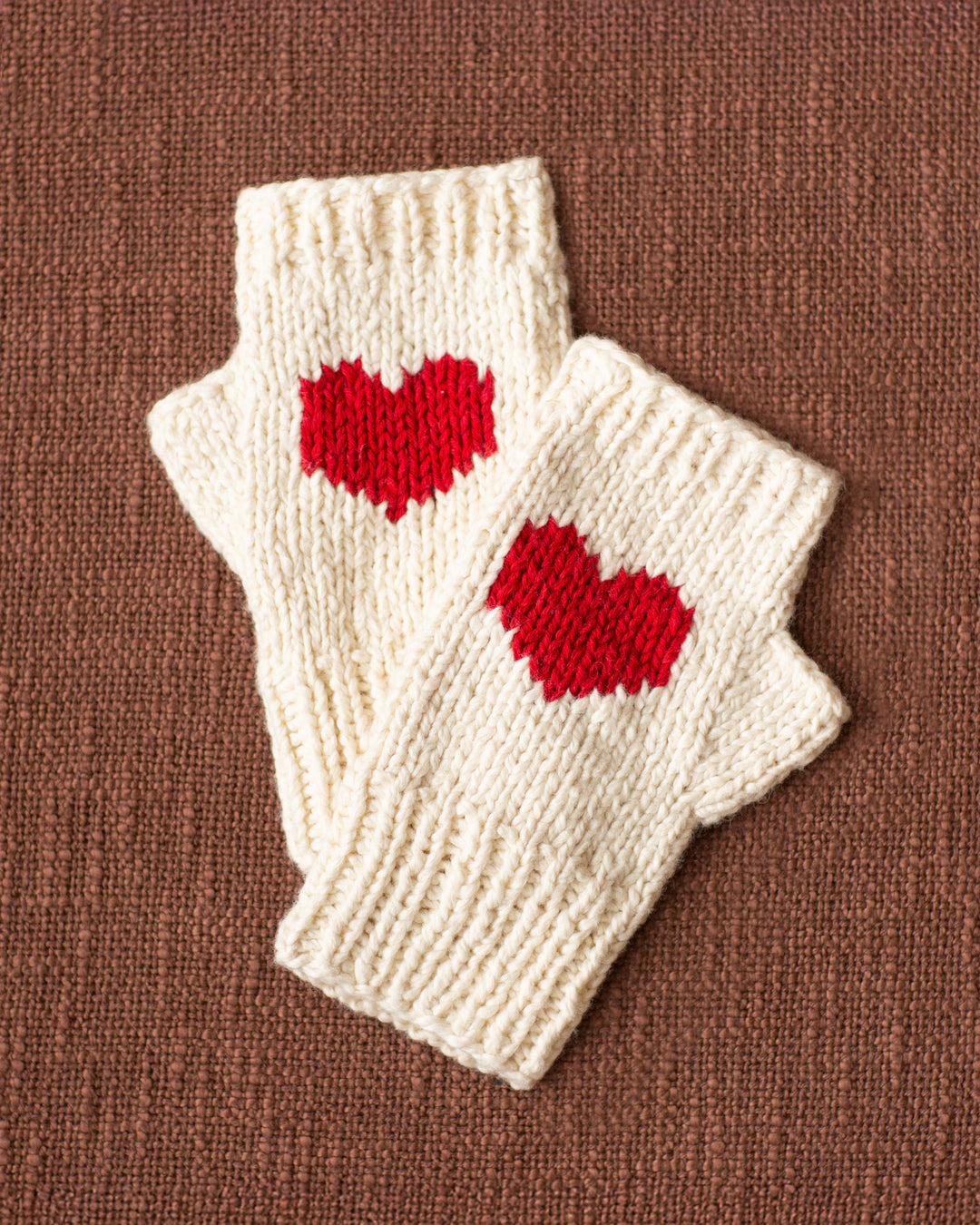 The Venus Heart Fingerless Glove features off-white, hand-knitted design with red heart patterns, displayed on a textured brown surface—an ideal cozy holiday gift.