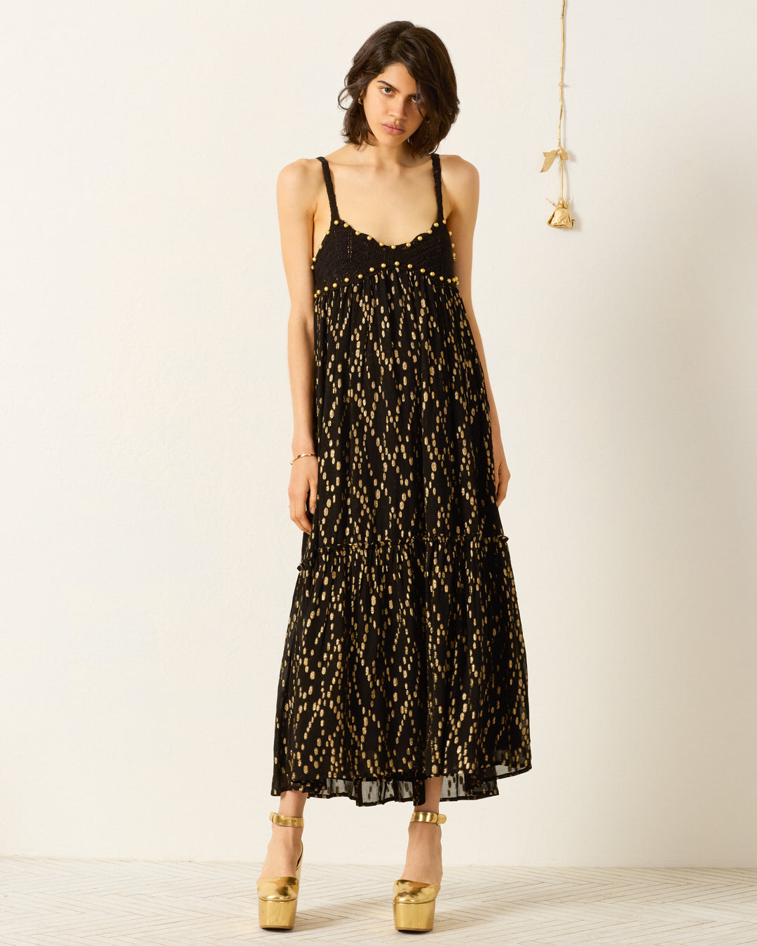 A woman in the Victoria Twilight Mist Dress, a handmade black maxi with gold fil coupé patterns, and gold platform heels stands against a plain white wall with hanging decor.