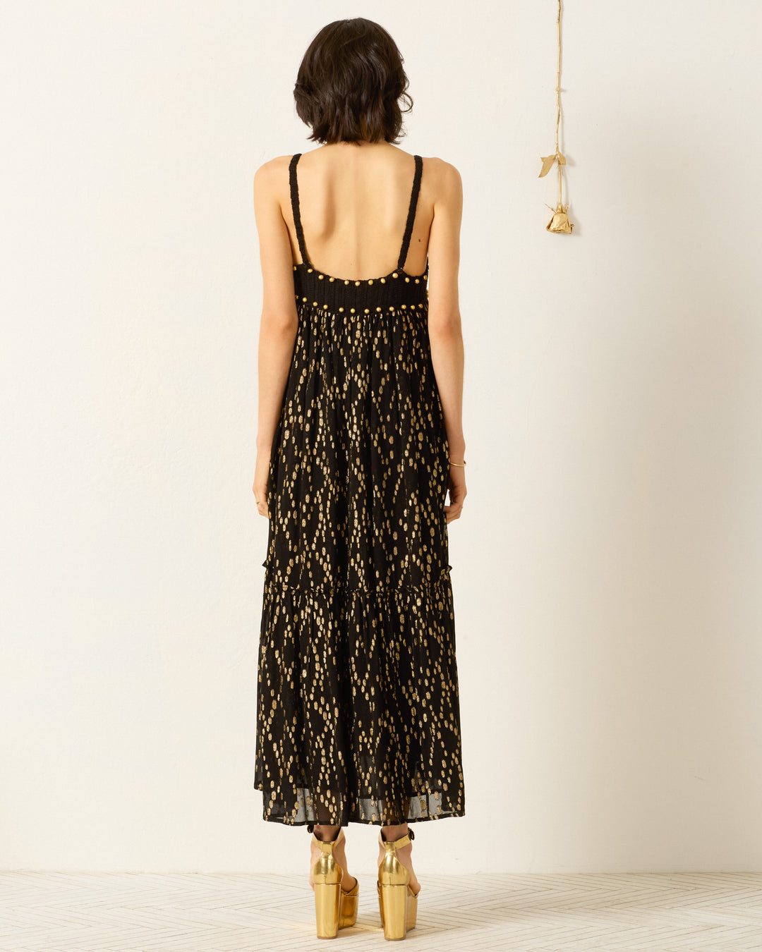 A woman faces away, wearing the Victoria Twilight Mist Dress—a sleeveless black empire-waist dress with gold fil coupé patterns—and platform sandals, standing against a plain light wall with a decorative hanging on the right.