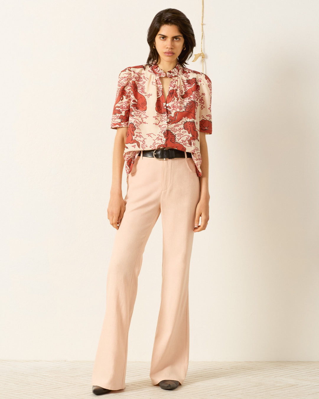 Person standing against a plain wall, wearing the Winnie Rouge Pegasus Shirt with puff shoulders, paired with beige high-waisted flared pants, a black belt, and black shoes.