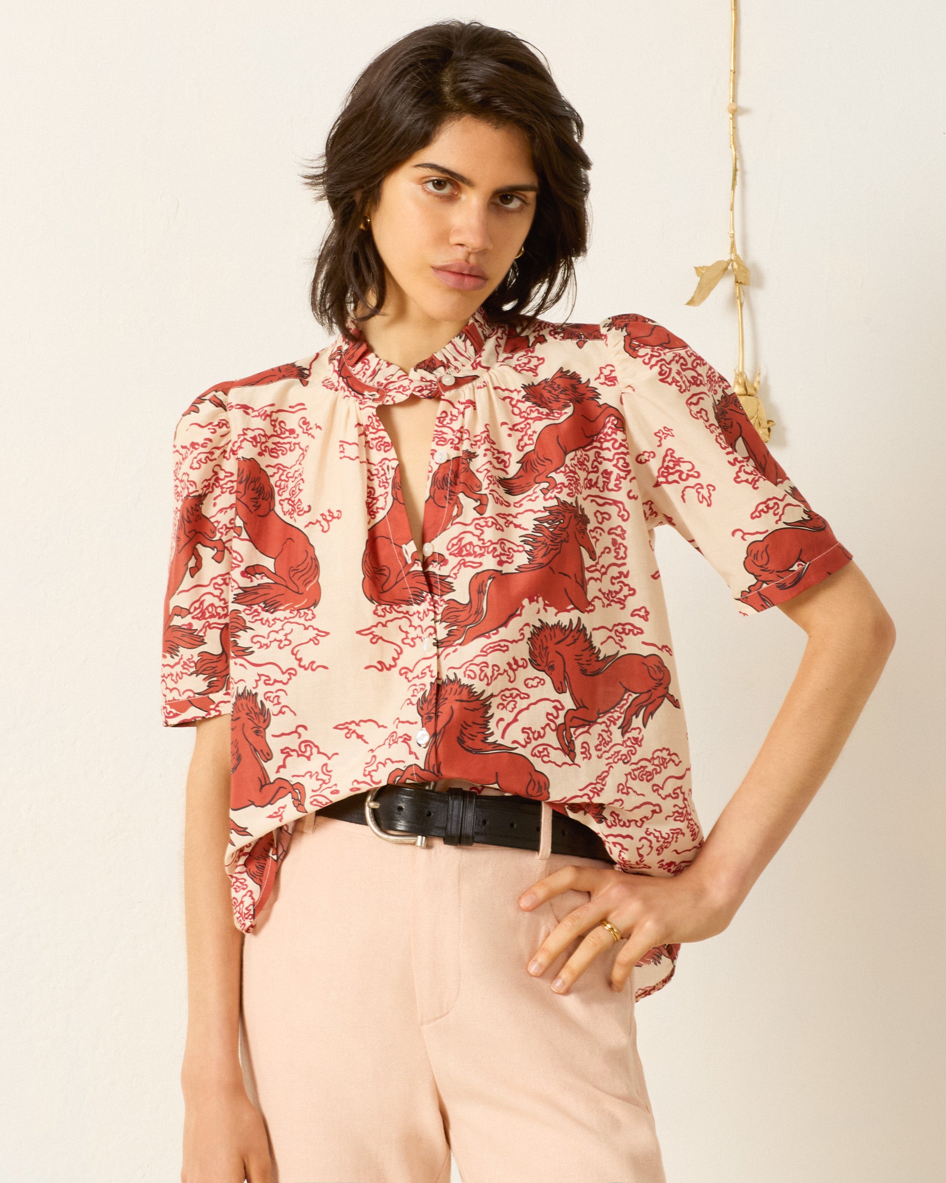 A woman stands against a plain off-white wall, wearing the Winnie Rouge Pegasus Shirt—a cream cotton-silk top with red horse print and puff shoulders—paired with light pink trousers and a black belt.