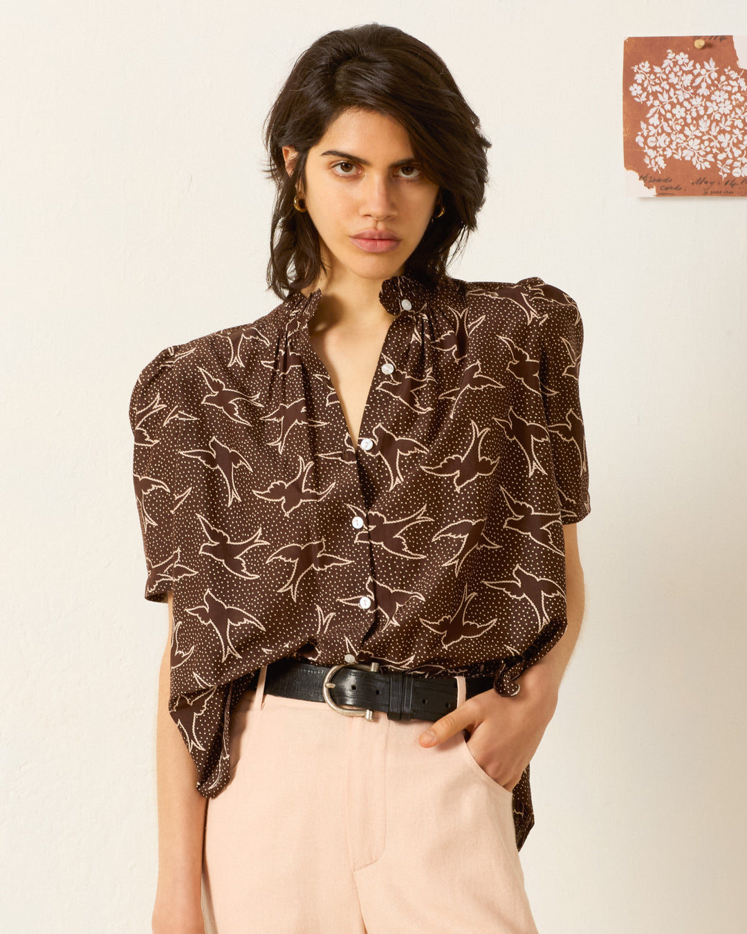 A woman wears the Winnie Clove Swallow Shirt, a brown blouse with white swallow prints, puff shoulders, and a ruffled collar, styled with pale pink pants and a black belt against a plain backdrop with art on the wall.