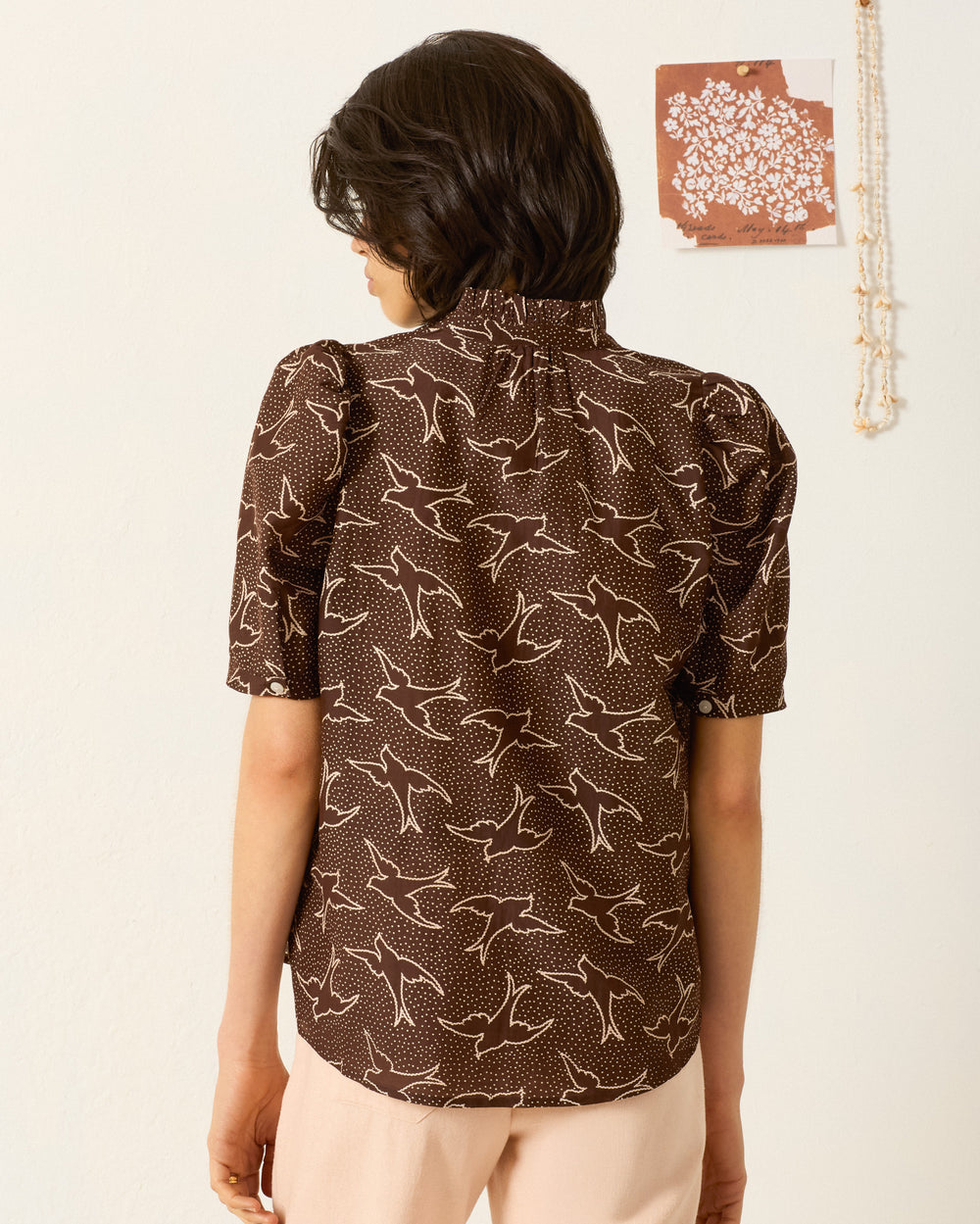A person facing away wears the Winnie Clove Swallow Shirt—a brown blouse with a white bird pattern and puff shoulders—paired with light pants, standing by a wall decorated with patterned art and a hanging beaded necklace.
