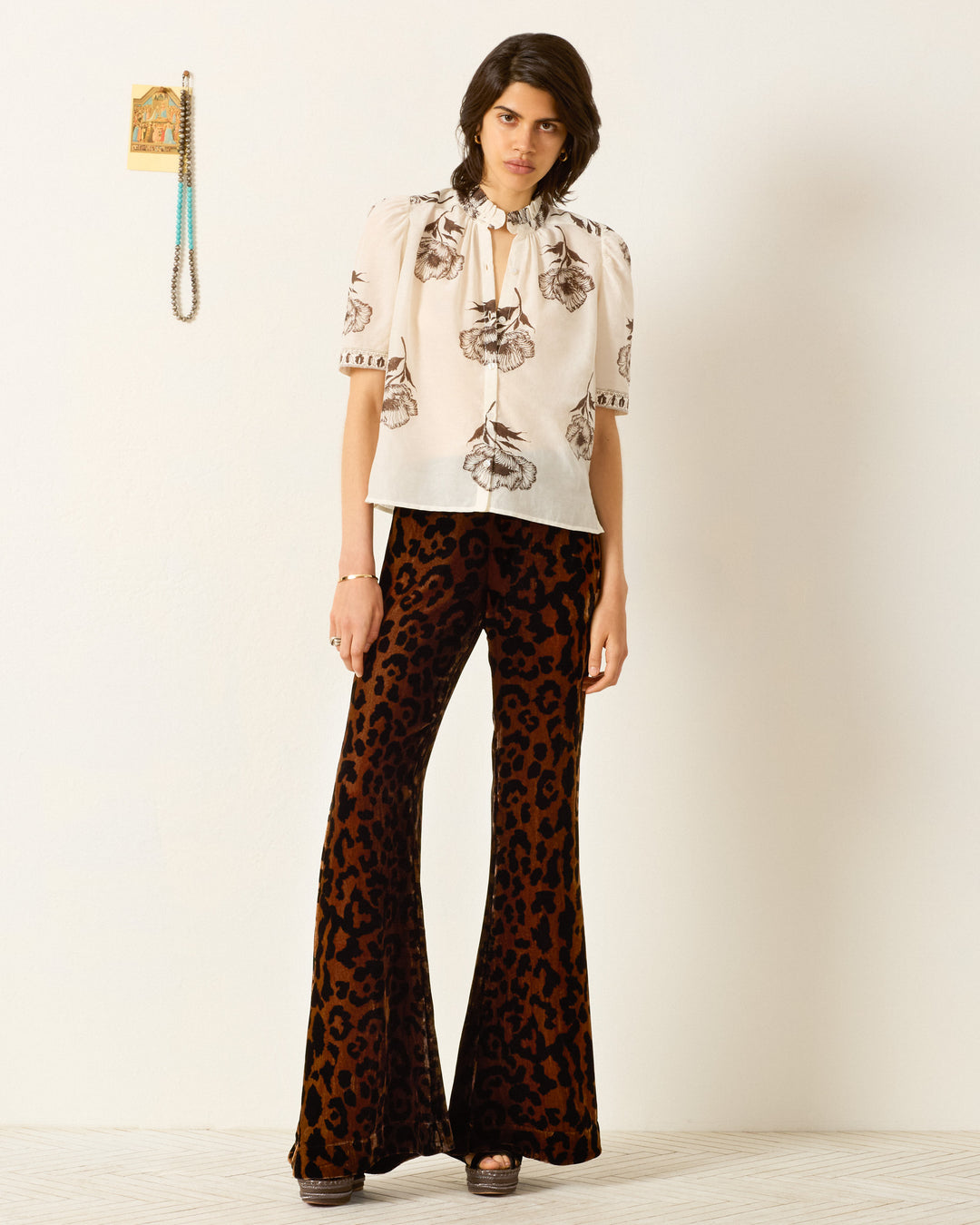 Indoors, a person wears the Winn Obsidian Poppy Shirt, a white cotton-silk blend top with floral prints, paired with high-waisted, brown and black leopard print flared pants.
