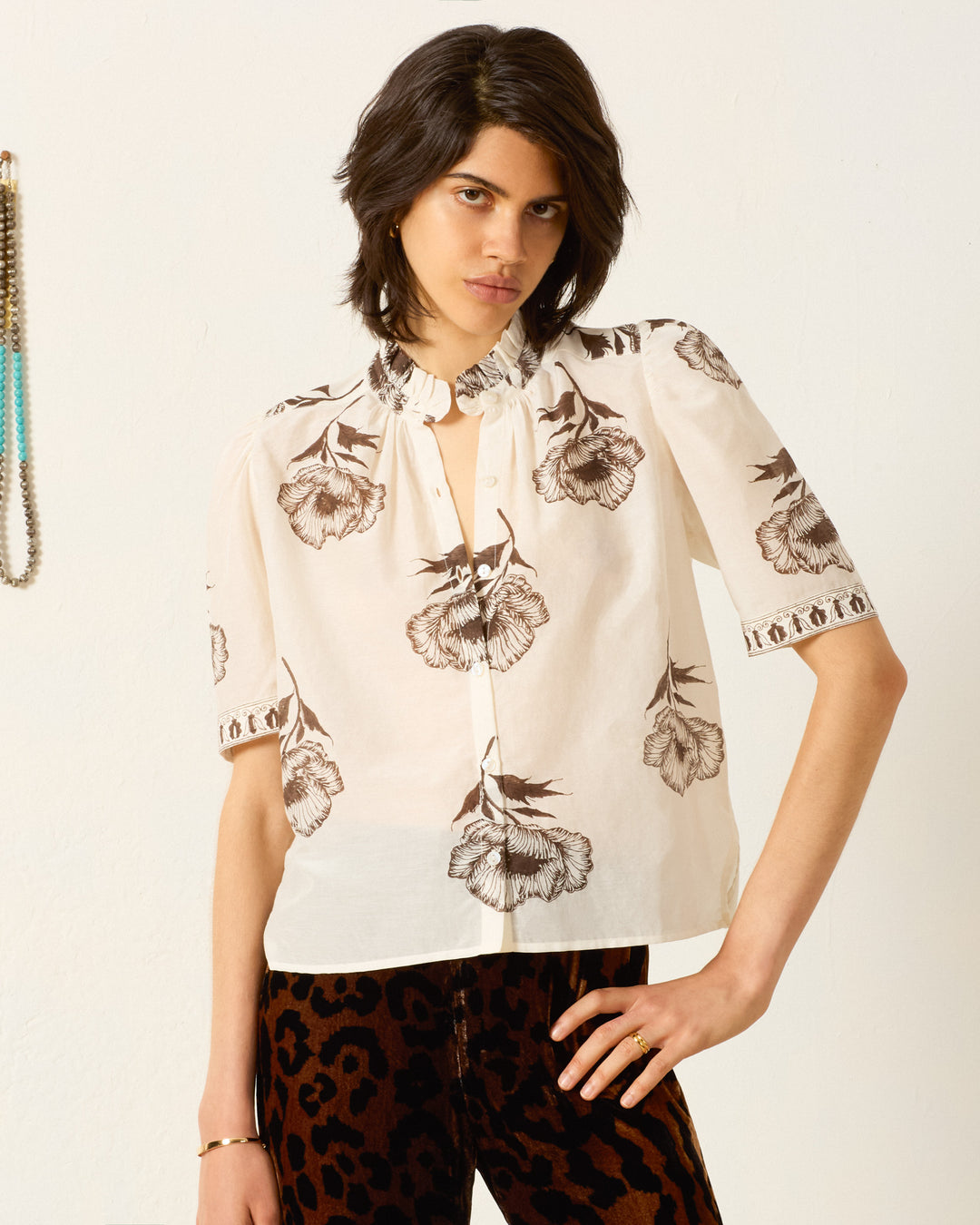 Someone stands against a plain, light wall wearing the Winn Obsidian Poppy Shirt—a white short-sleeve cotton-silk blend blouse with a black poppy motif—paired with high-waisted animal print pants.