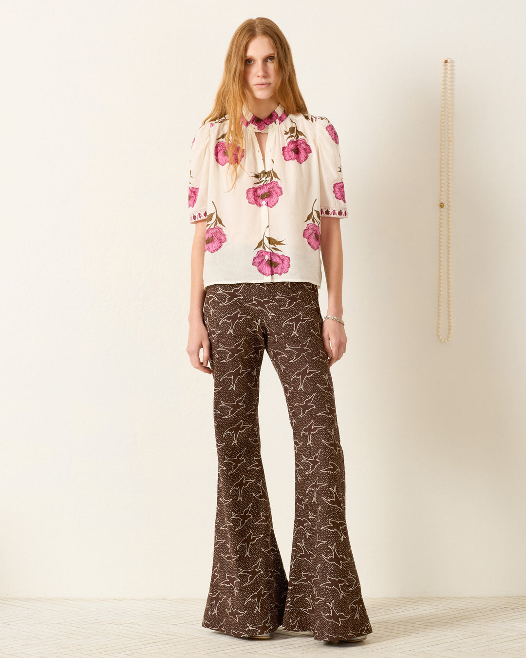 A woman stands indoors wearing the Winn Fuchsia Poppy Shirt—a hand-block-printed white blouse with pink floral prints—and brown flared pants with a white pattern; a long beaded necklace hangs on the wall behind her.