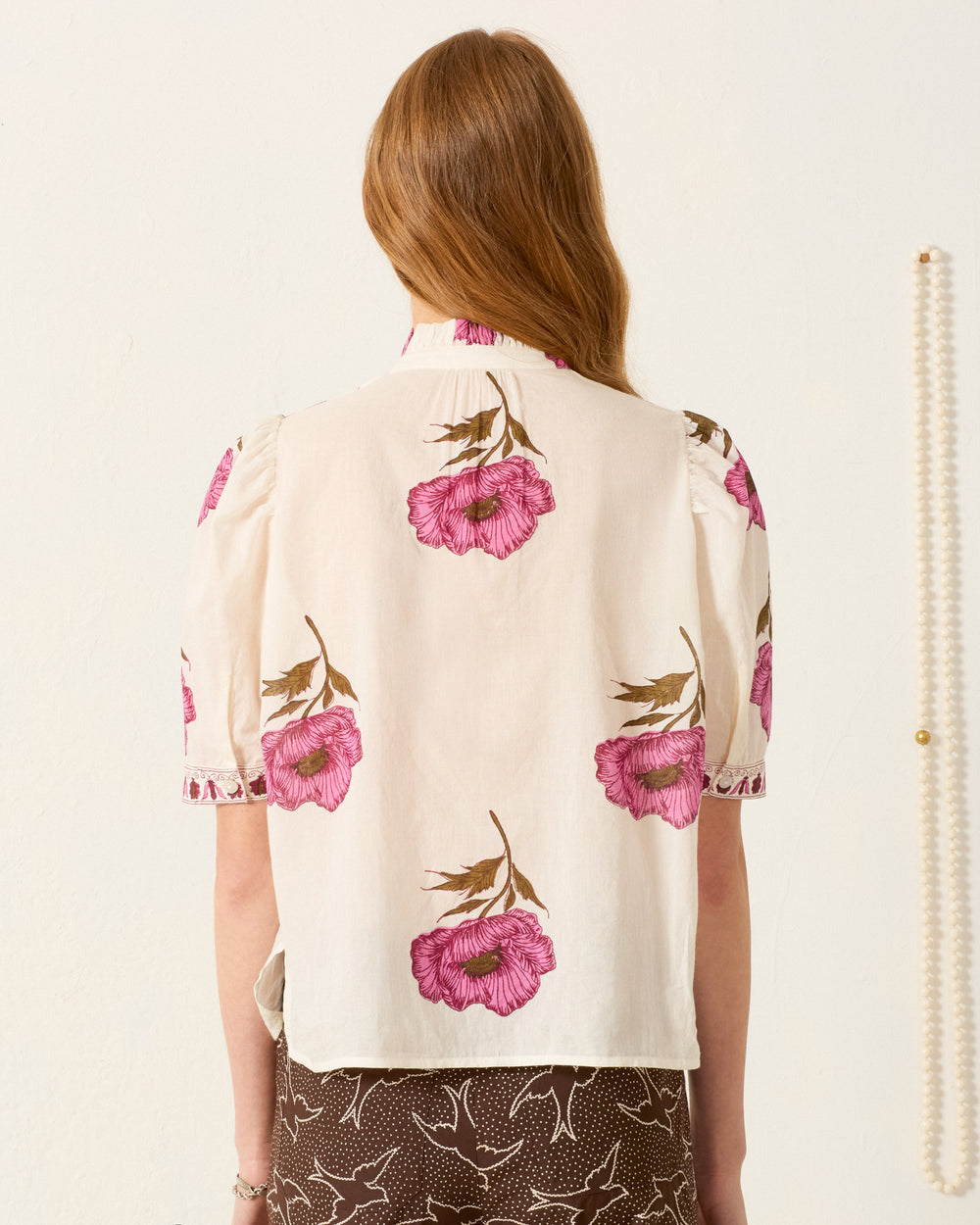 A woman with long hair is seen from behind wearing the Winn Fuchsia Poppy Shirt, a hand-block-printed white short-sleeve blouse with pink floral prints, paired with a brown skirt featuring a white bird pattern.