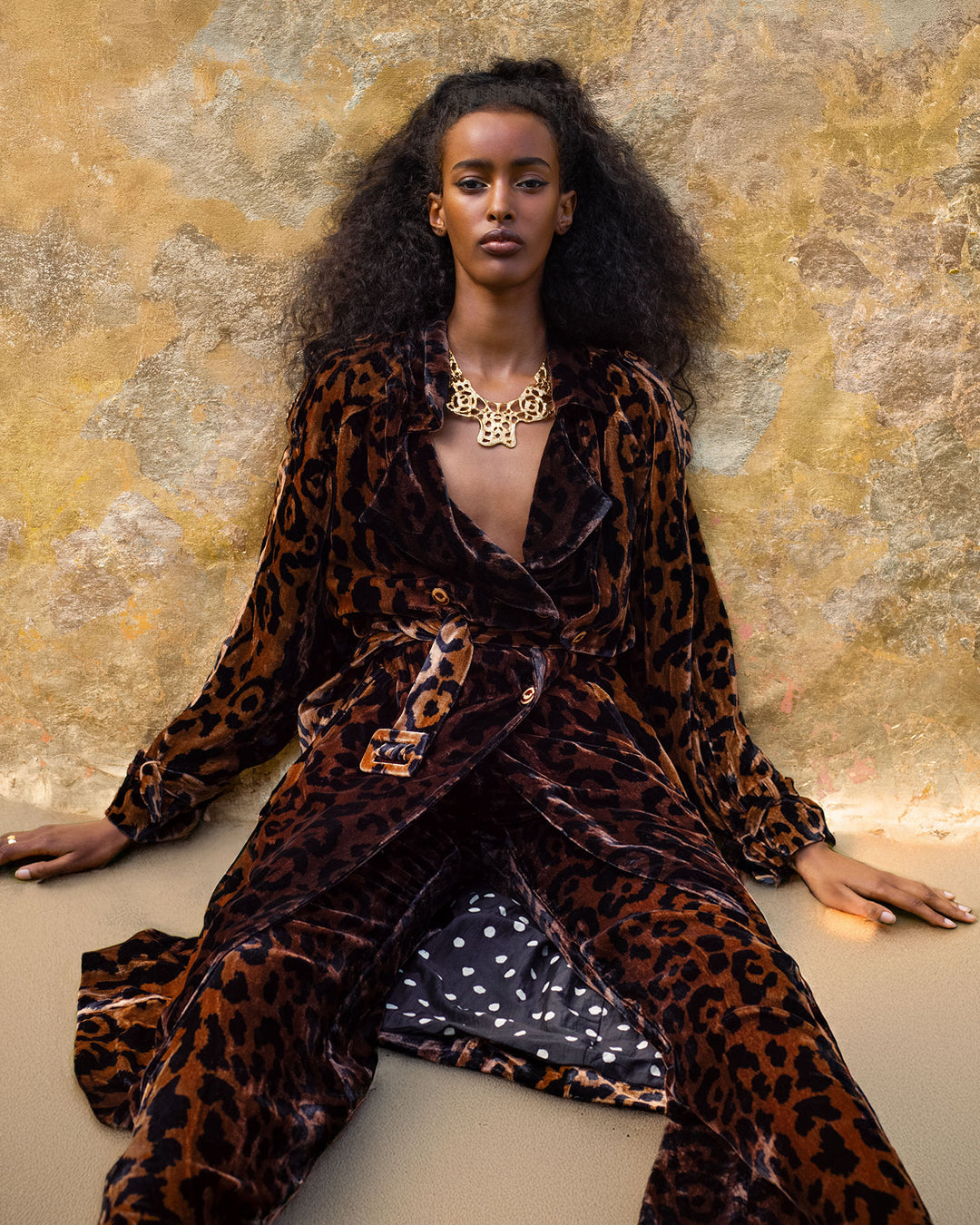 A woman poses against a textured wall, wearing the Jasper Leopard Velvet Coat, styled with a gold statement necklace and loose, voluminous hair.